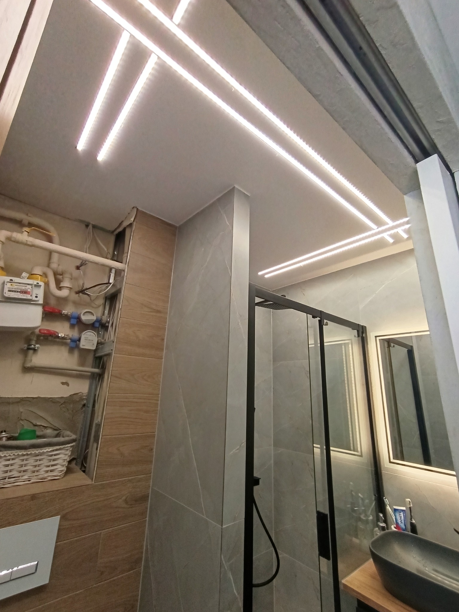 Wykończona łazienka z kabiną prysznicową, umywalką na blacie i ledowym oświetleniem sufitowym. Widoczne rury i liczniki przed zabudową.