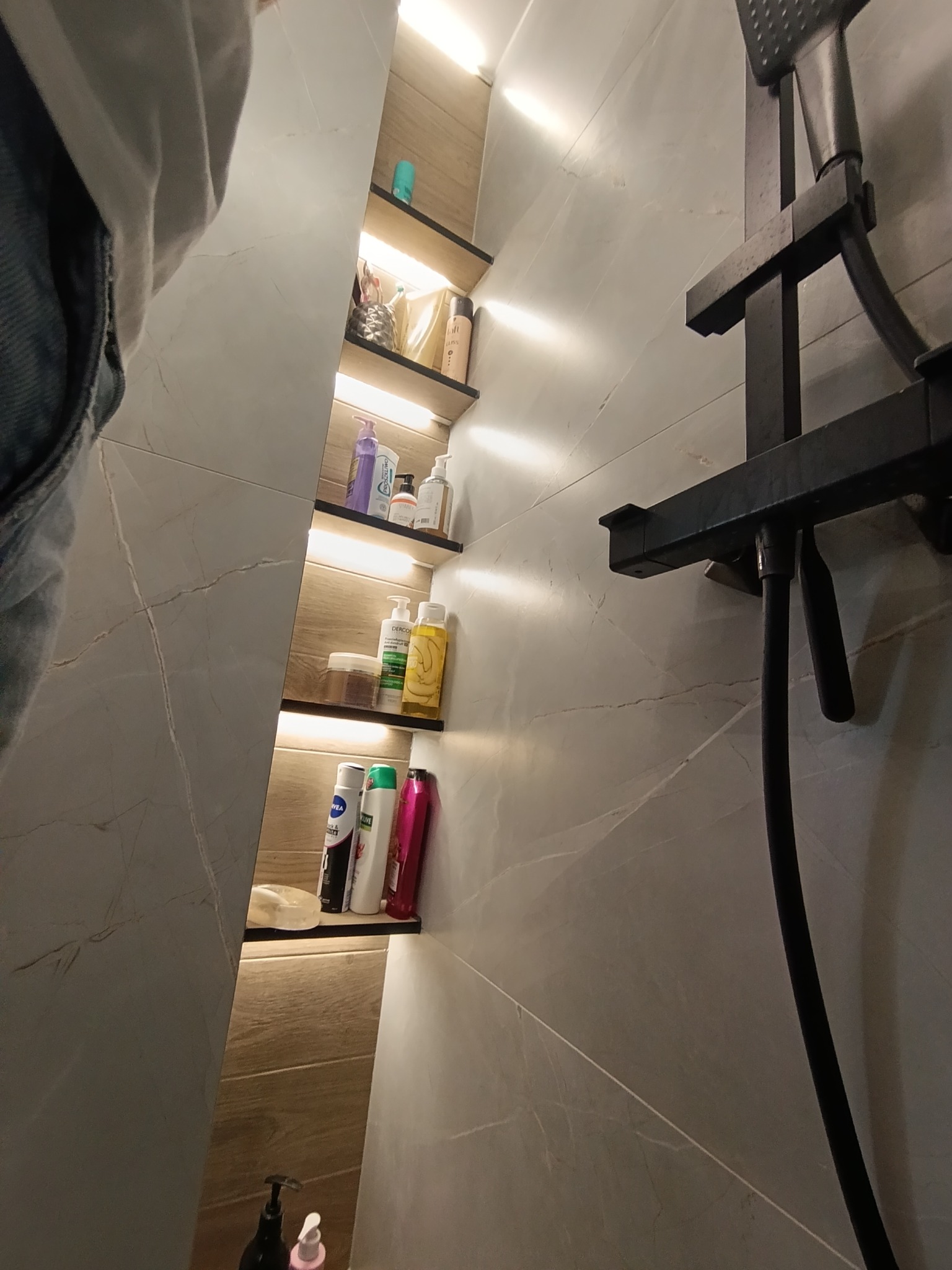 Nowoczesna łazienka z wnęką oświetloną LED, wypełnioną kosmetykami. Widoczny fragment kabiny prysznicowej z czarną armaturą i marmurowe płytki ścienne.