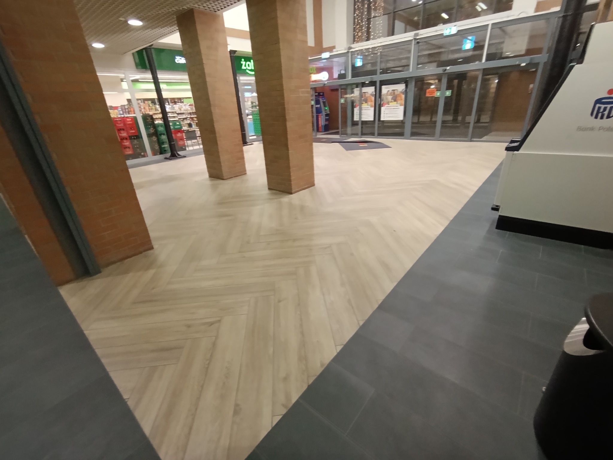 Jasna, drewnopodobna podłoga ułożona w jodełkę w przestrzeni komercyjnej, kontrastująca z ciemnymi płytkami. Widoczne filary i wejście do sklepu.