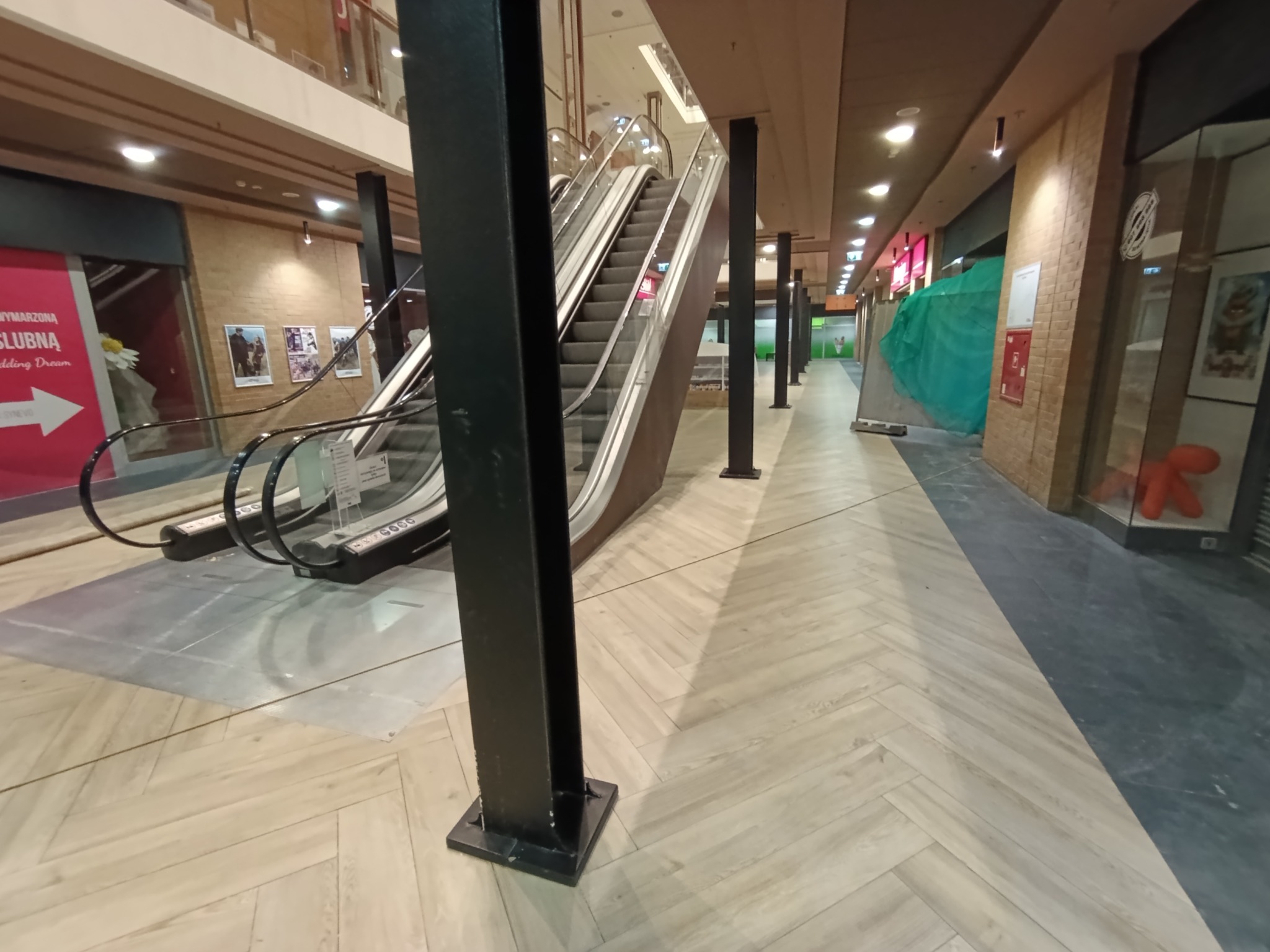 Jasna, drewnopodobna podłoga ułożona w jodełkę w pasażu handlowym, widoczny fragment schodów ruchomych i czarne słupy konstrukcyjne.