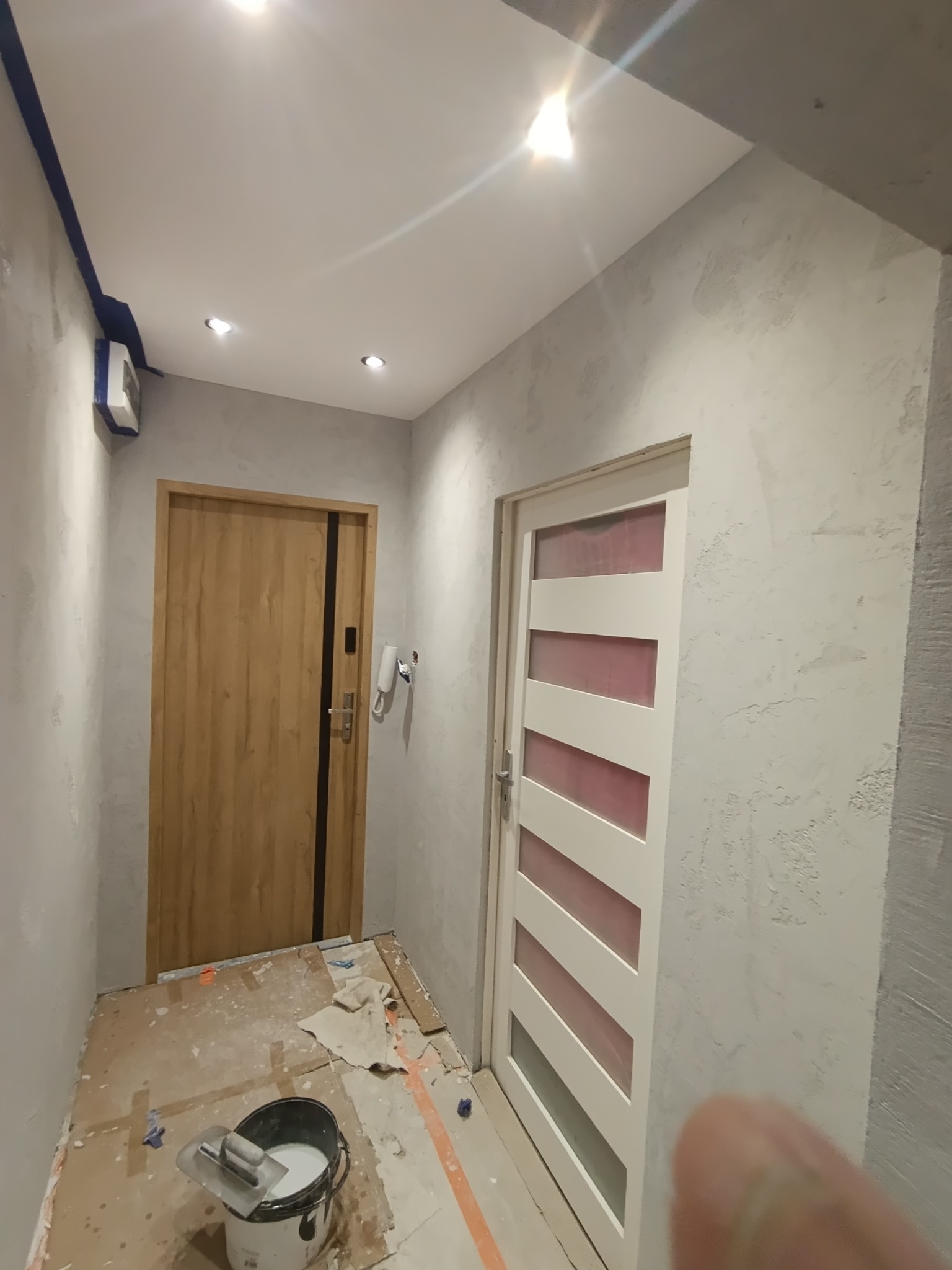 Korytarz w trakcie prac: ściany pokryte szarą strukturą, widoczne dwie różne ościeżnice drzwiowe, zabezpieczona podłoga i wiadro z narzędziem.