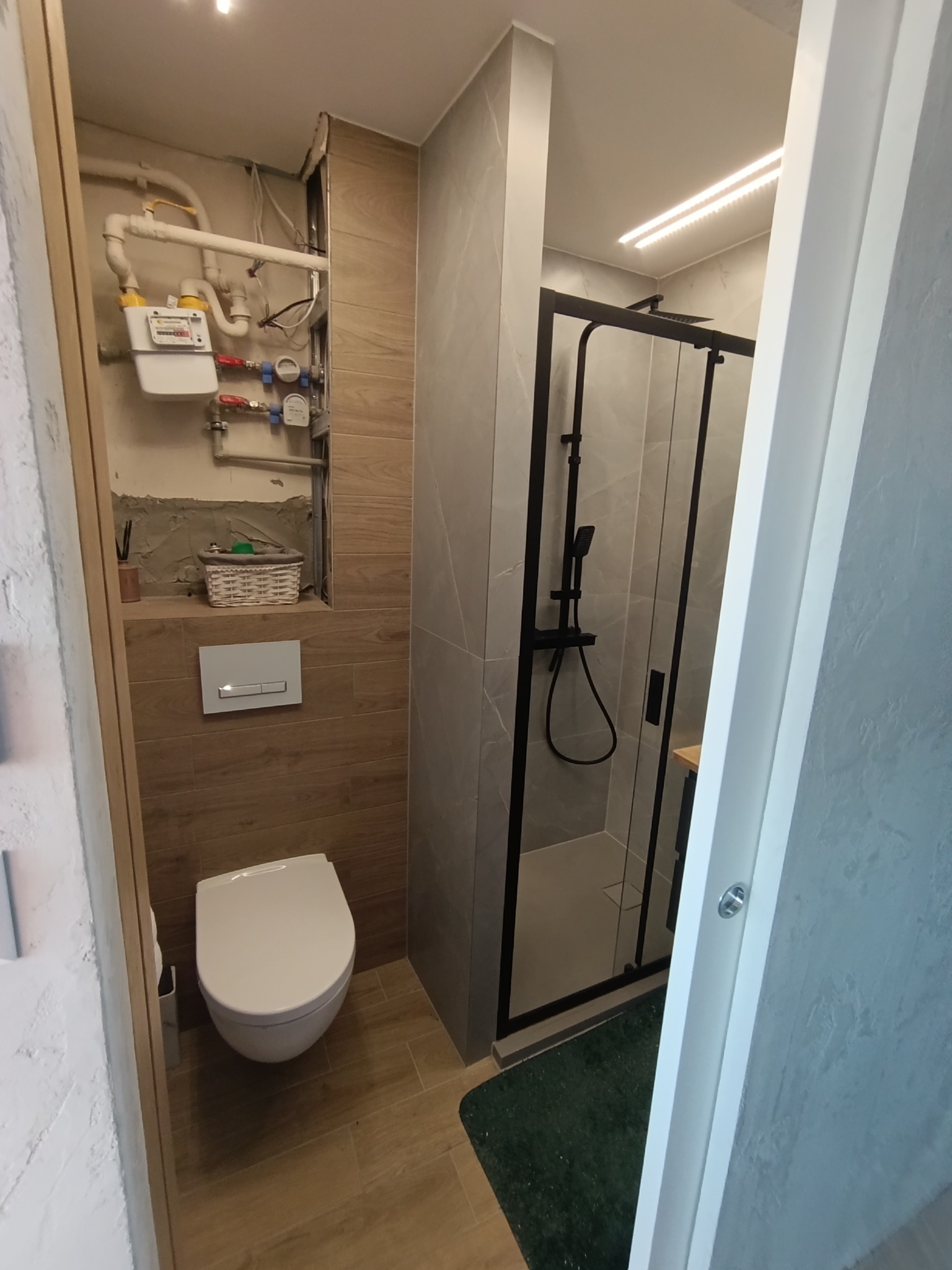 Nowoczesna łazienka z kabiną prysznicową typu walk-in, wiszącą toaletą i drewnopodobnymi płytkami. Widoczne piony wod-kan i gazowe.