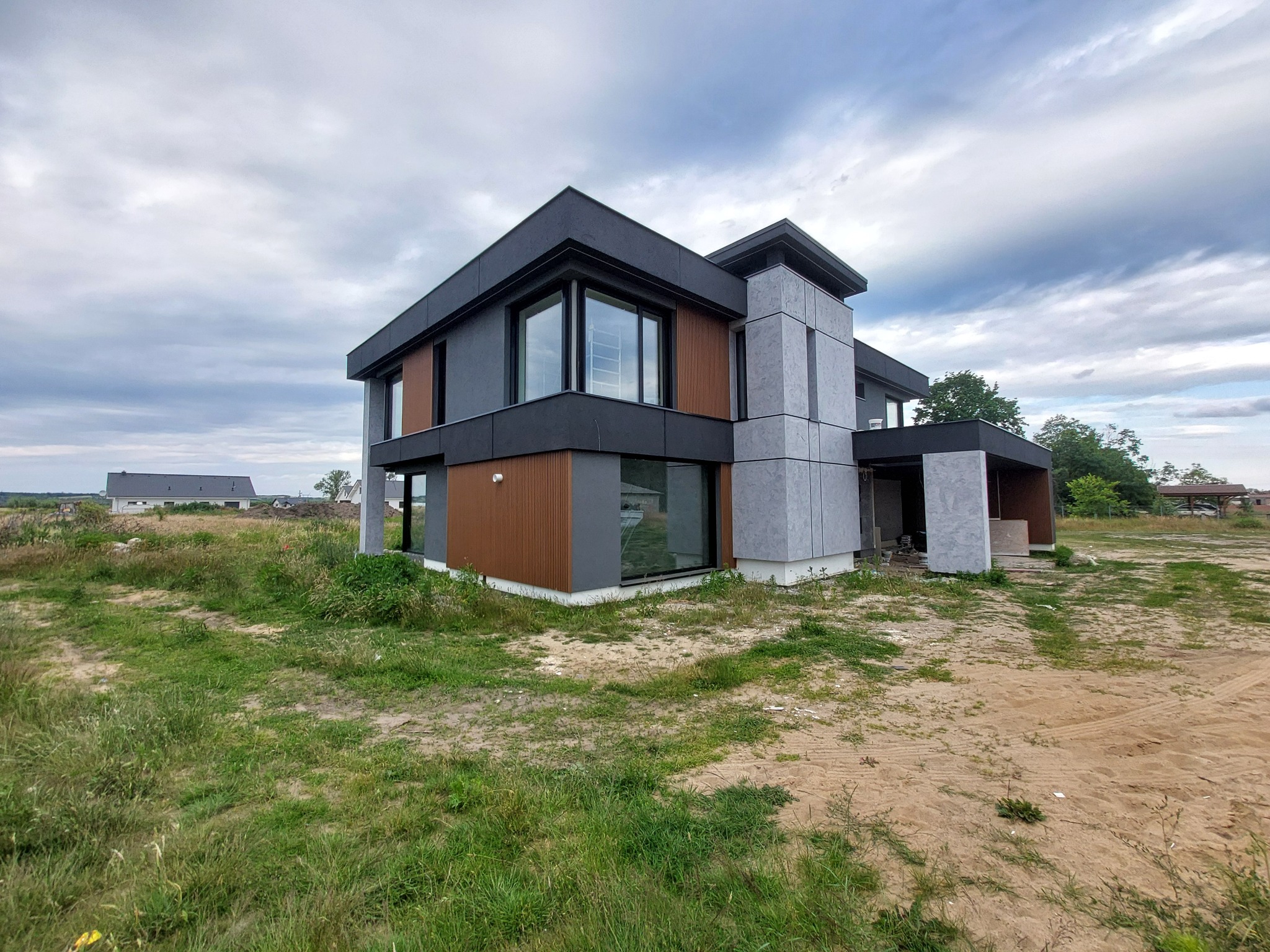 Nowoczesny dom z elewacją z paneli w odcieniach szarości i brązu, duże okna, otoczenie zarośnięte trawą. Widok z dołu, pochmurne niebo.