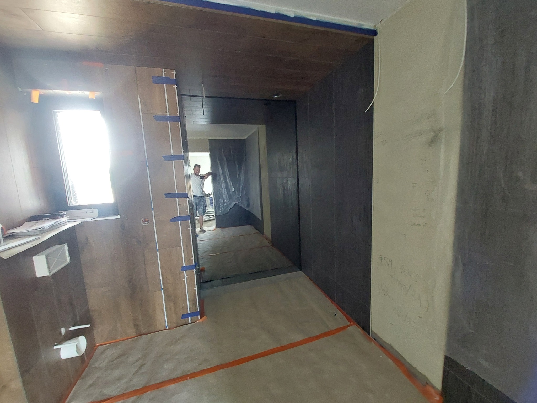 Wnętrze w trakcie prac: ściany wyłożone płytami, widoczny fragment instalacji zabezpieczony taśmą. W tle postać w przejściu, podłoga zabezpieczona folią.
