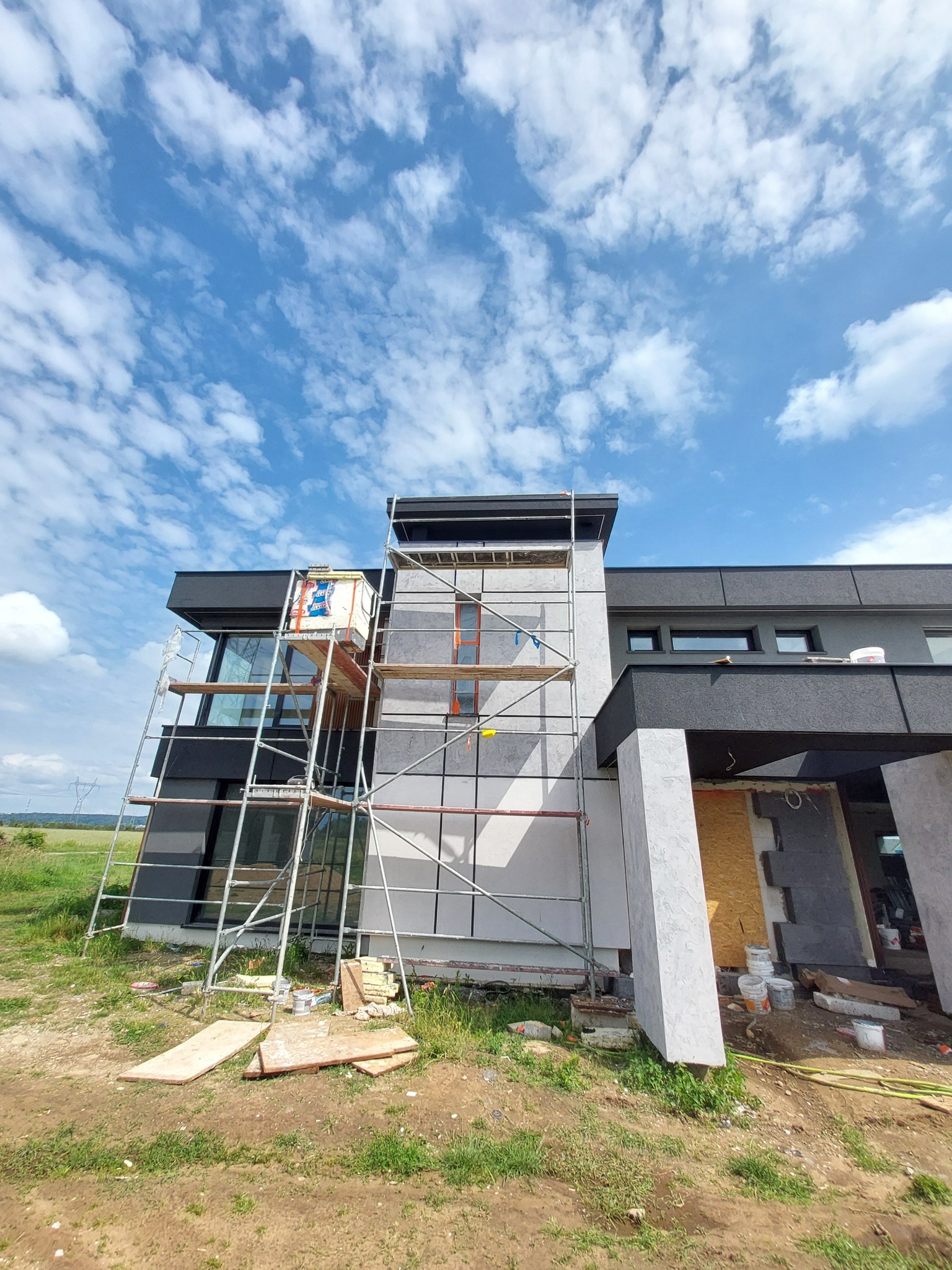 Elewacja domu w trakcie wykończenia z rusztowaniem na tle błękitnego nieba z chmurami. Widoczne materiały budowlane i elementy dekoracyjne.
