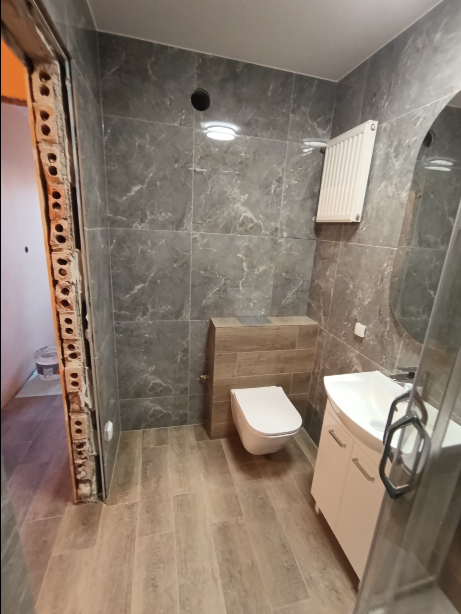 Wykończona łazienka z szarymi płytkami imitującymi marmur, drewnopodobną zabudową WC i białymi meblami. Widoczny fragment surowej ściany przy wejściu.