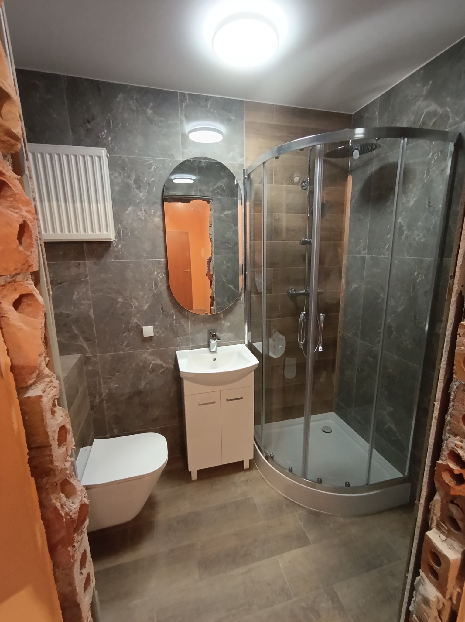 Ukończona łazienka z szarymi płytkami, kabiną prysznicową, umywalką z szafką i toaletą. Fragment ściany z cegły w tle dodaje surowego charakteru.