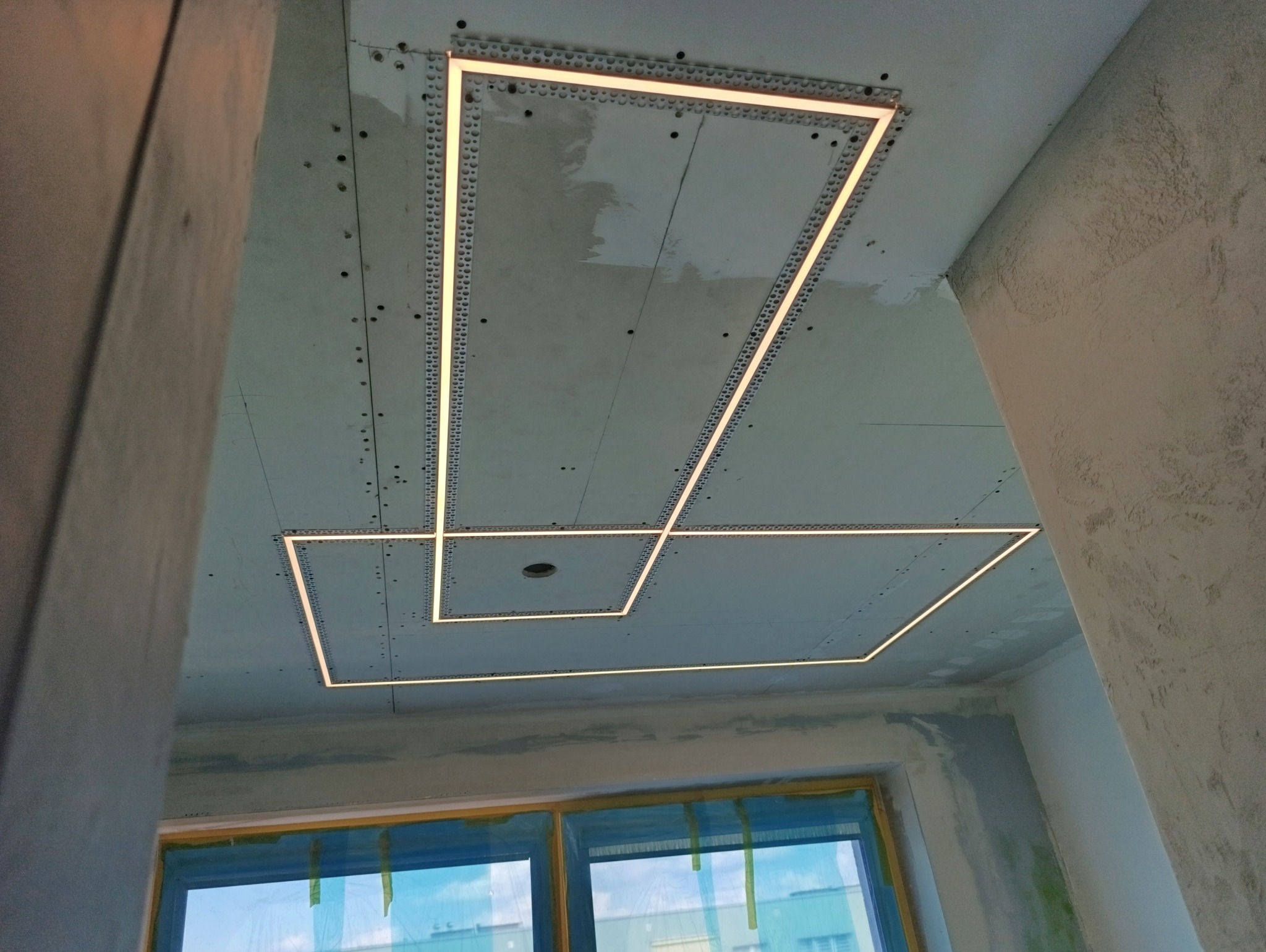 Sufit z płyt gipsowo-kartonowych z zamontowanym oświetleniem LED w kształcie kwadratów, widoczne łączenia płyt i szpachlowanie. W tle okno z folią ochronną.