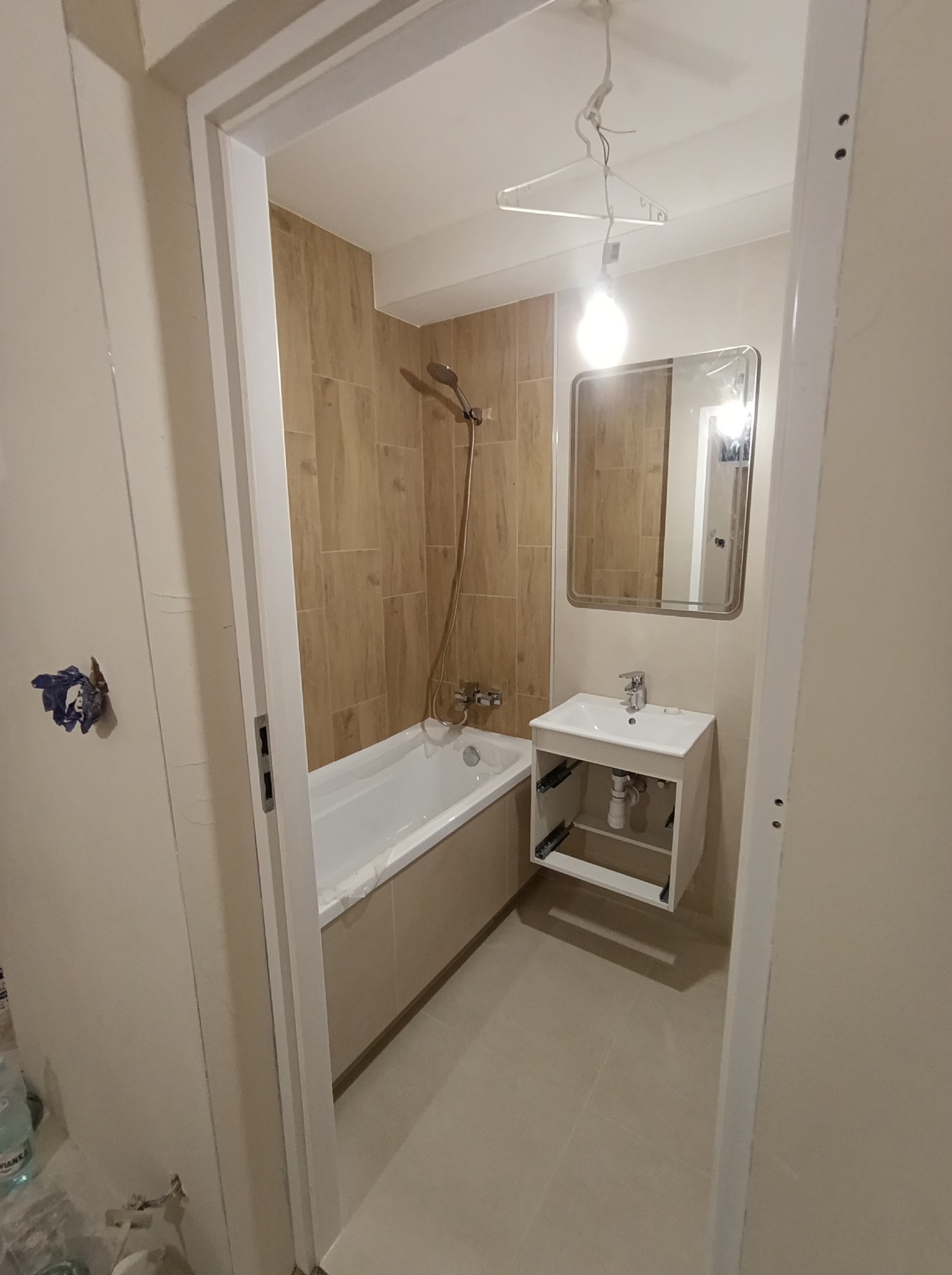 Jasna łazienka po remoncie: beżowe płytki imitujące drewno na ścianach, biała wanna i umywalka z szafką, lustro. Widok zza futryny drzwi.