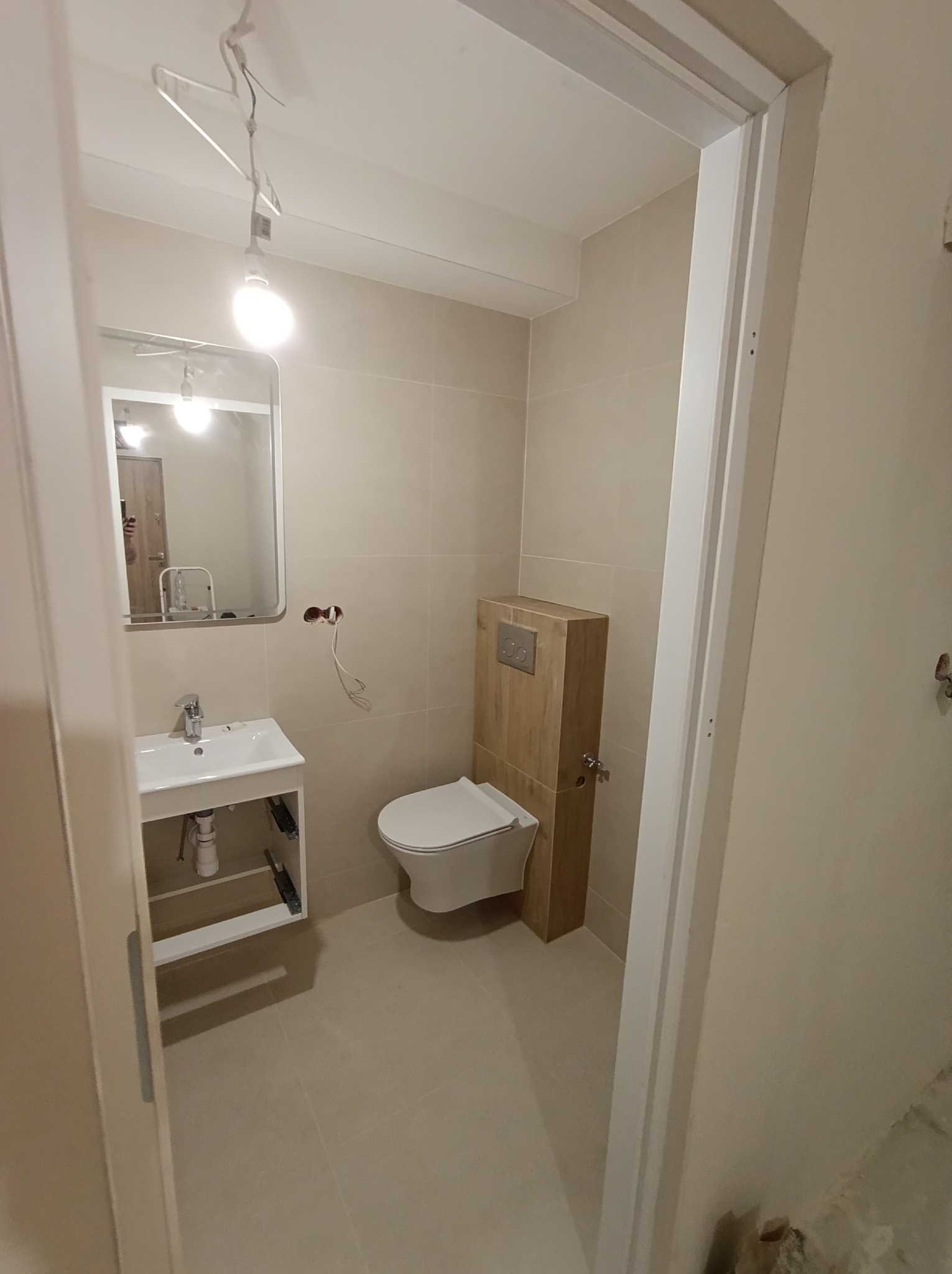 Jasna łazienka w trakcie wykańczania: beżowe płytki, umywalka z szafką, toaleta z zabudowaną spłuczką. Widoczne niedokończone elementy elektryczne.