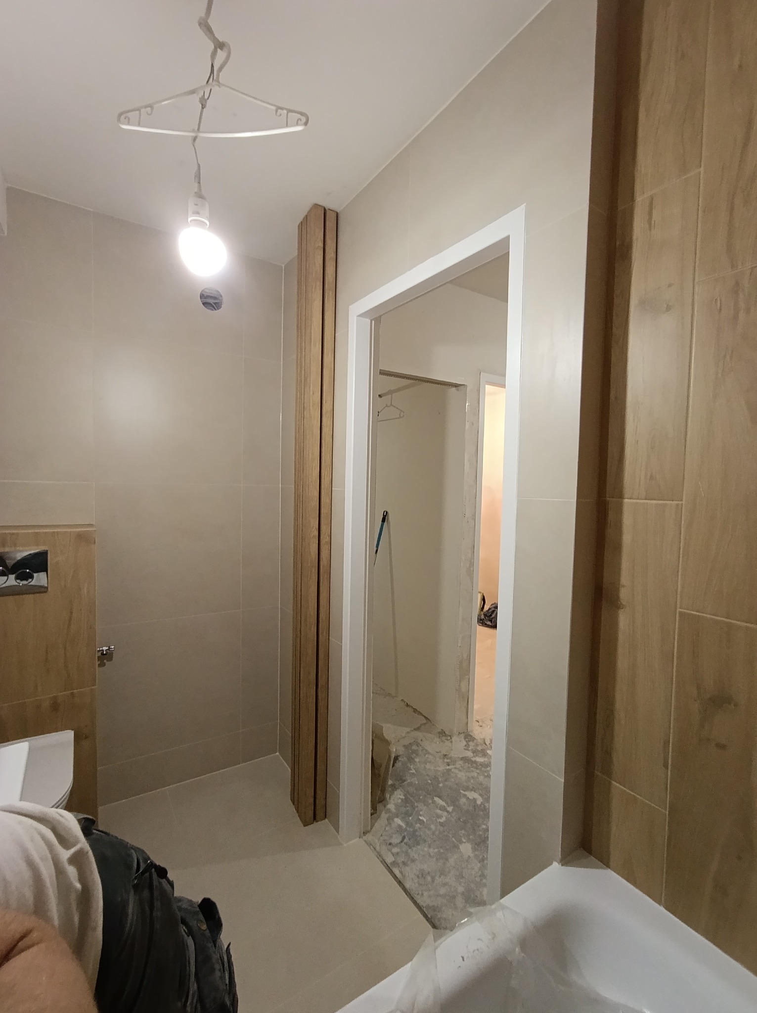 Jasna łazienka w trakcie wykańczania: beżowe płytki, drewniane akcenty, biała armatura. Widoczny fragment wanny i wejście do kolejnego pomieszczenia.