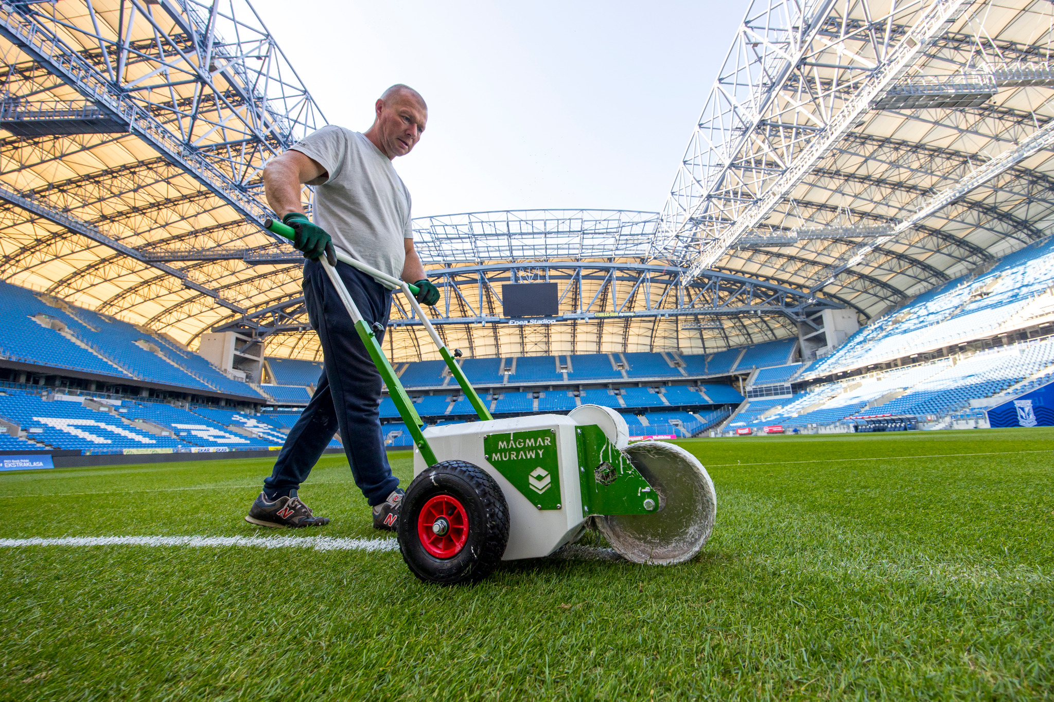 Pracownik z użyciem wózka do malowania linii boiska sportowego na tle trybun stadionu.