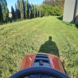ZałogaBruk - Kosiarka traktorek Husqvarna w trakcie koszenia trawnika, widoczny cień operatora, równo skoszona trawa, rząd drzew w tle, dom po prawej stronie.