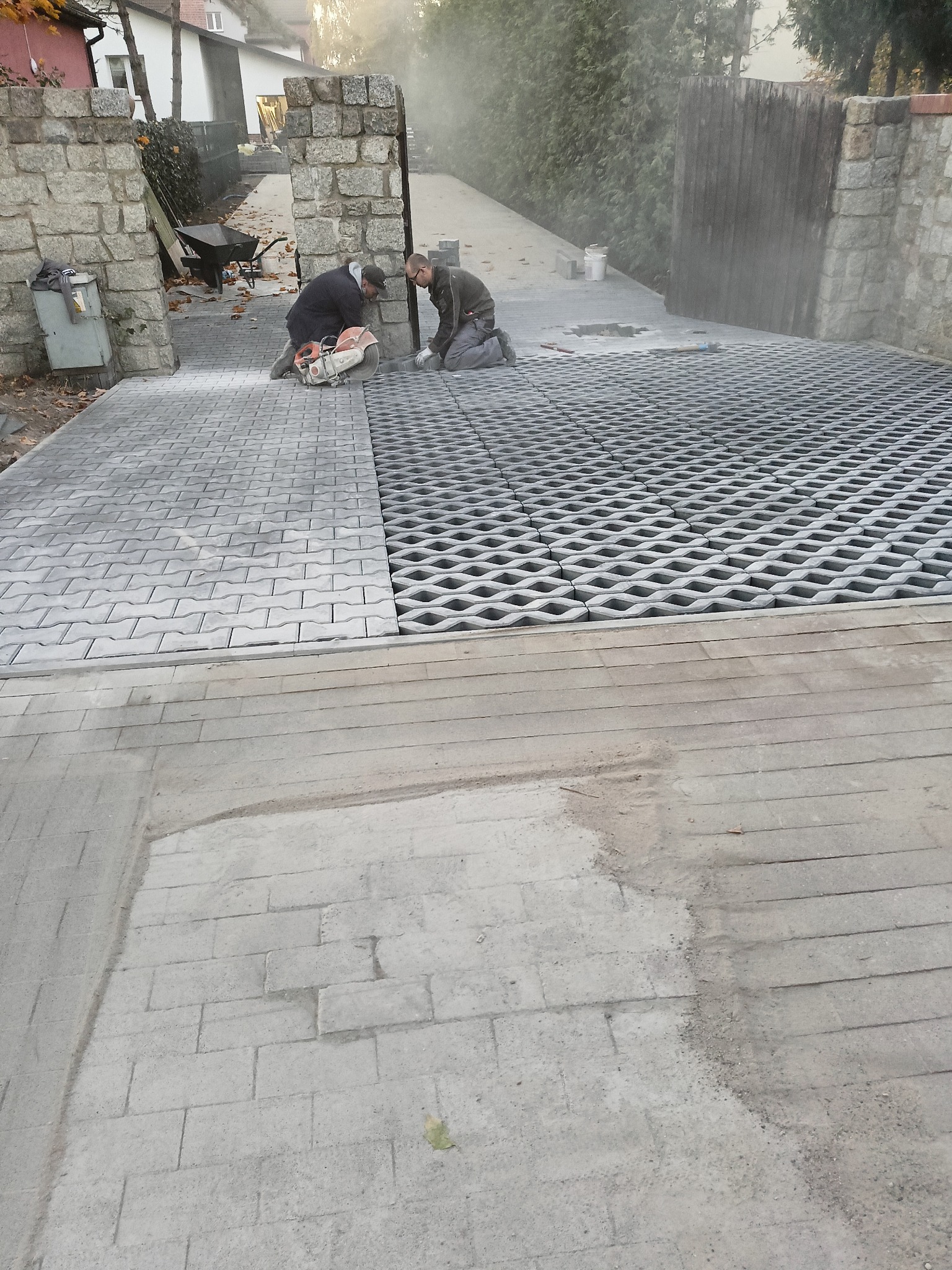 Dwóch pracowników przycina szarą kostkę brukową za pomocą piły spalinowej, układając ją na podjeździe z ażurowych płyt betonowych, widoczne ogrodzenie z kamienia łupanego i wózek budowlany w tle.
