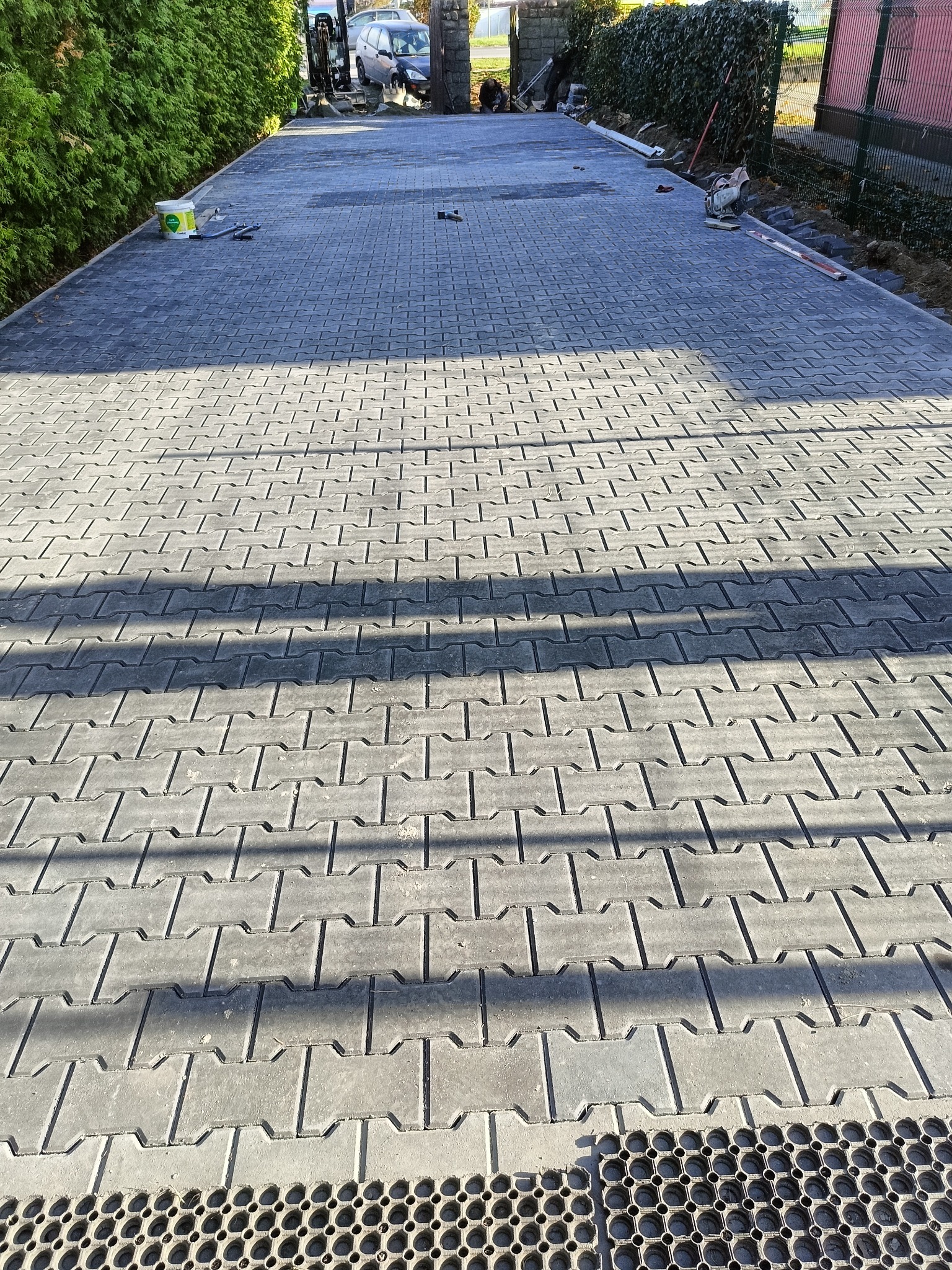 Ułożona szara kostka brukowa na podjeździe, widoczny fragment kratki ściekowej, w tle zaparkowany samochód i zieleń.