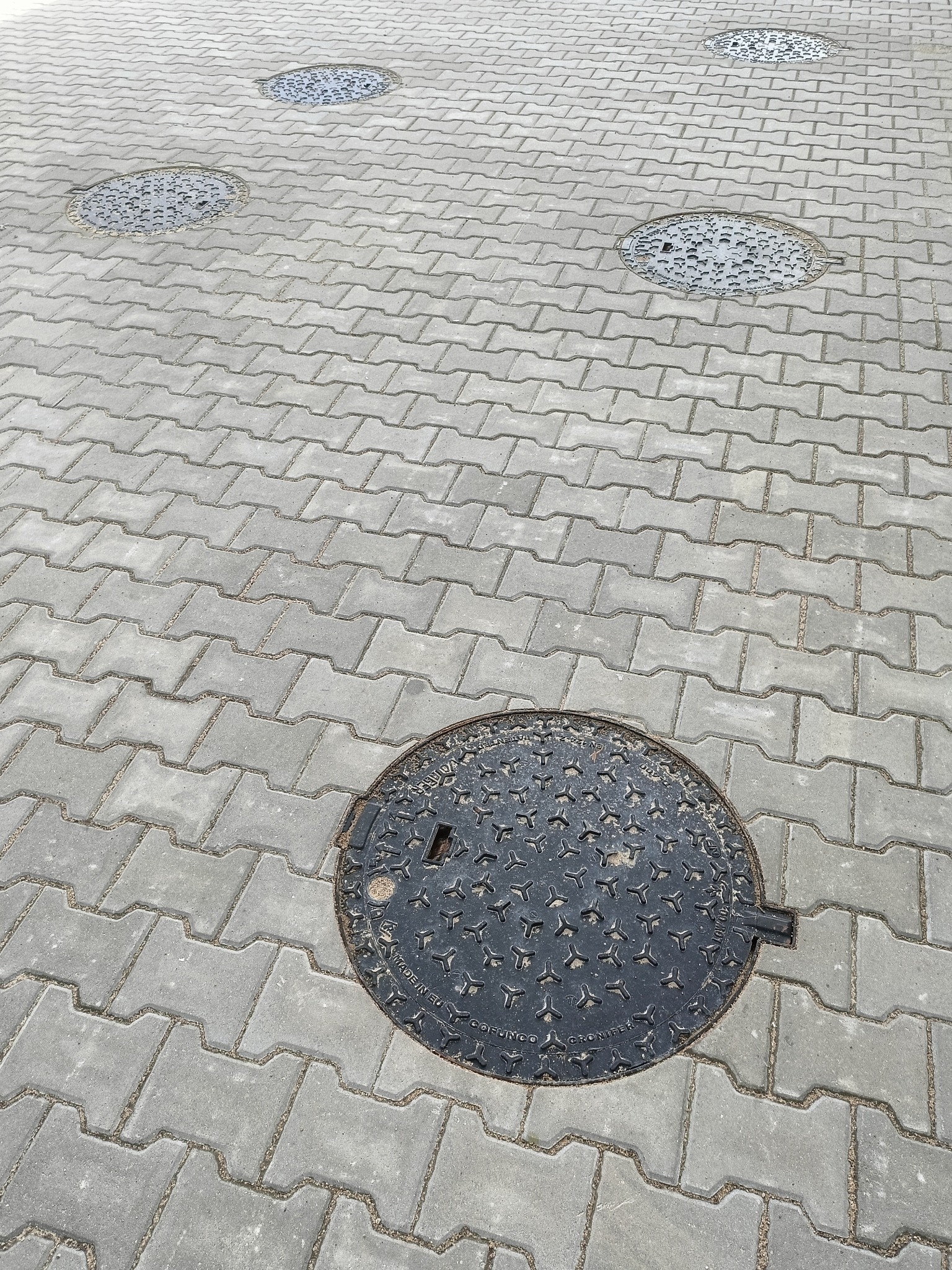 Szara kostka brukowa typu 'I' z czterema okrągłymi włazami kanalizacyjnymi z żeliwa w oddali.