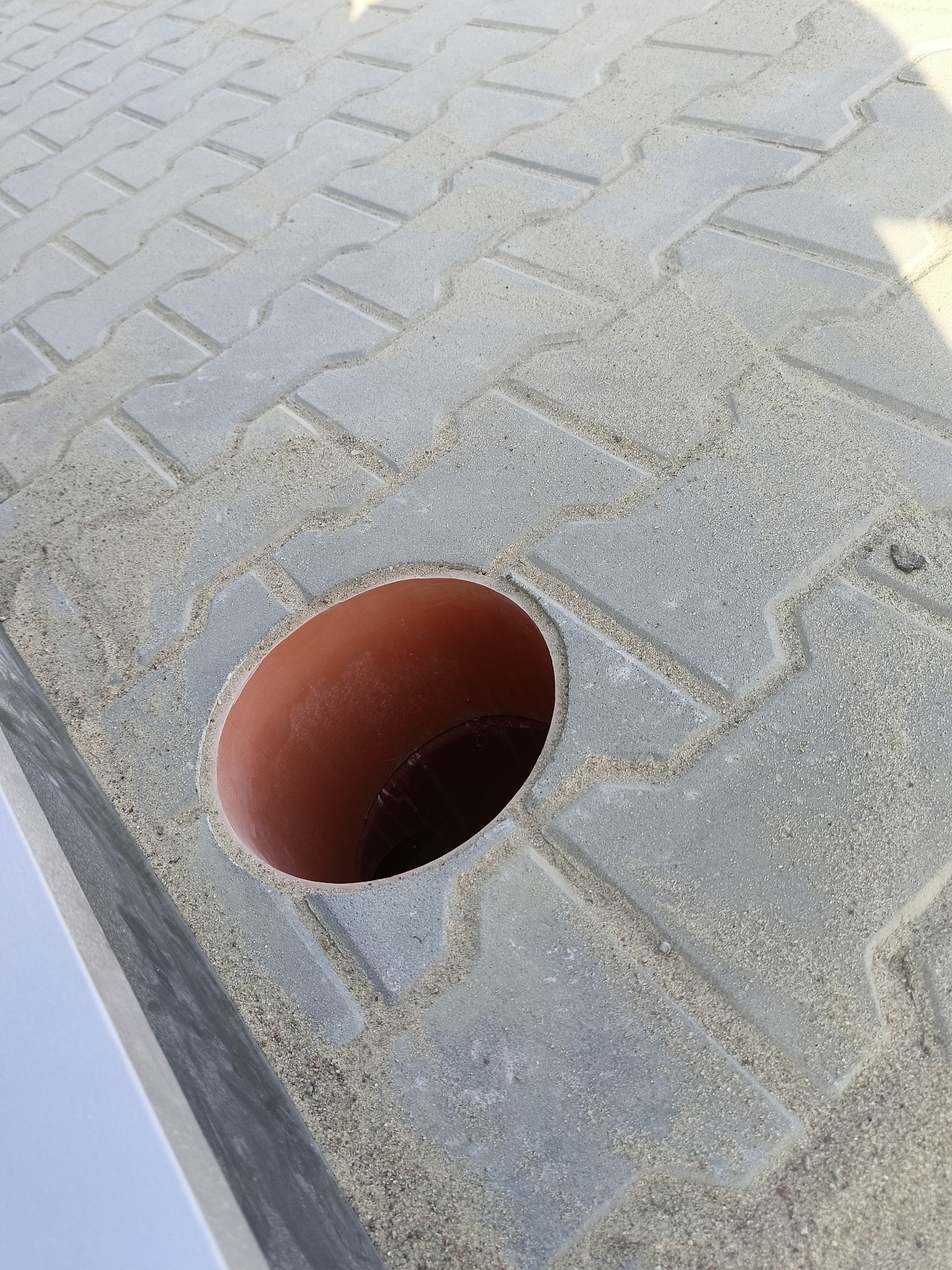 Szare kostki brukowe ułożone wokół pomarańczowej rury wystającej z ziemi, zasypane piaskiem.