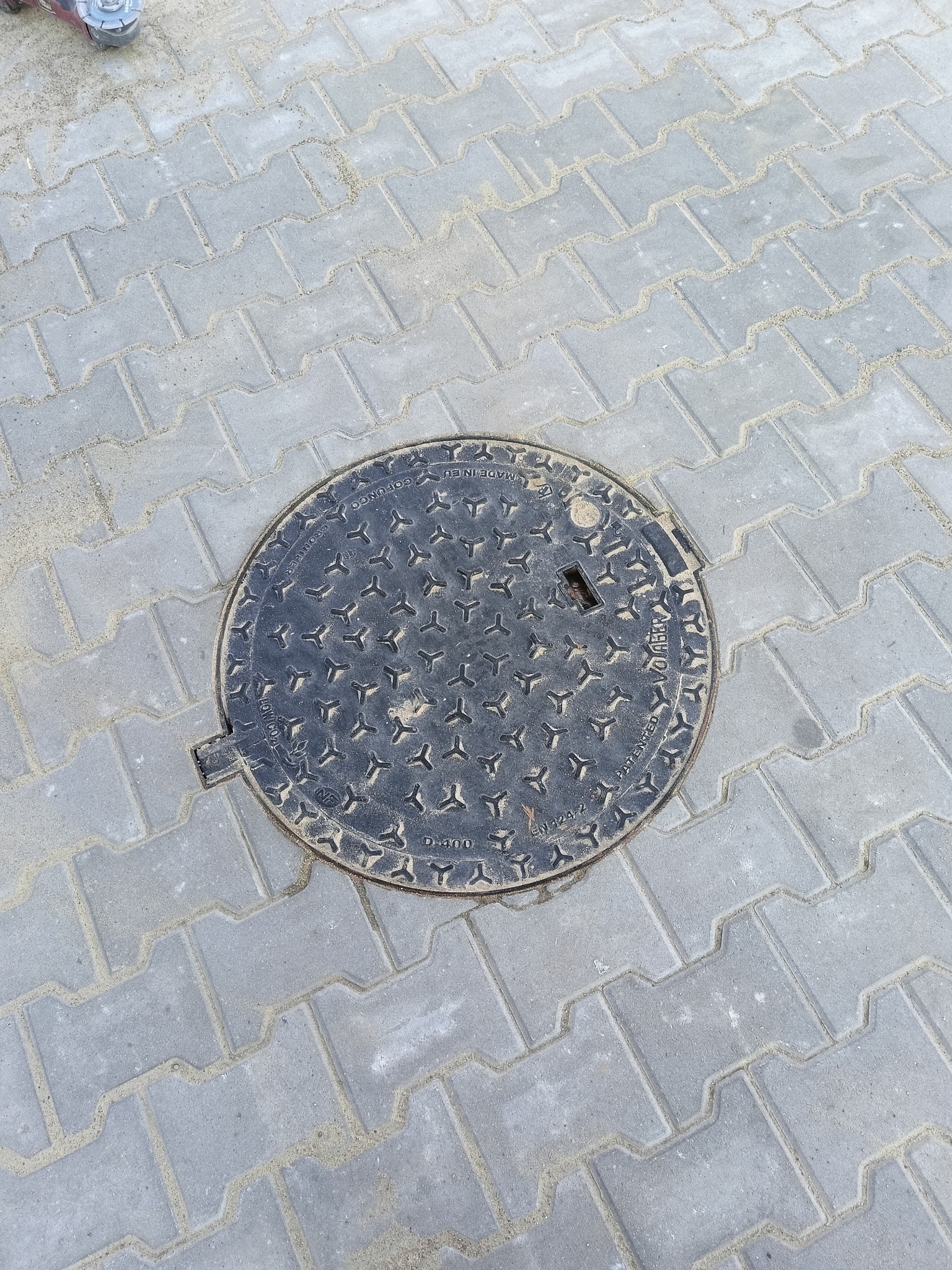 Szara kostka brukowa ułożona wokół okrągłej, żeliwnej studzienki kanalizacyjnej z widocznymi oznaczeniami producenta i wzorem antypoślizgowym.