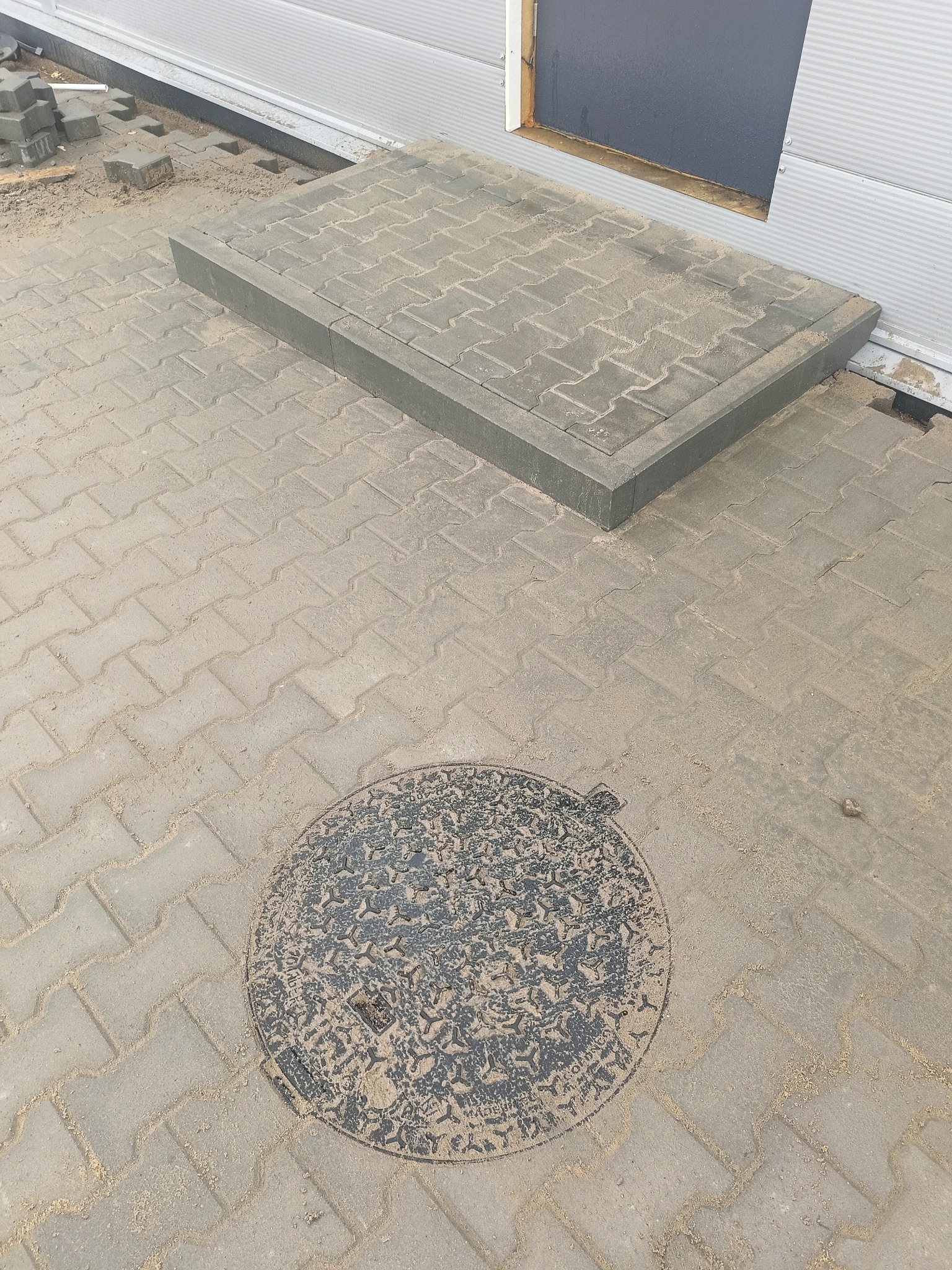 Ułożona kostka brukowa przed budynkiem z metalową elewacją, widoczny właz kanalizacyjny i niedokończone obrzeże schodów wejściowych.