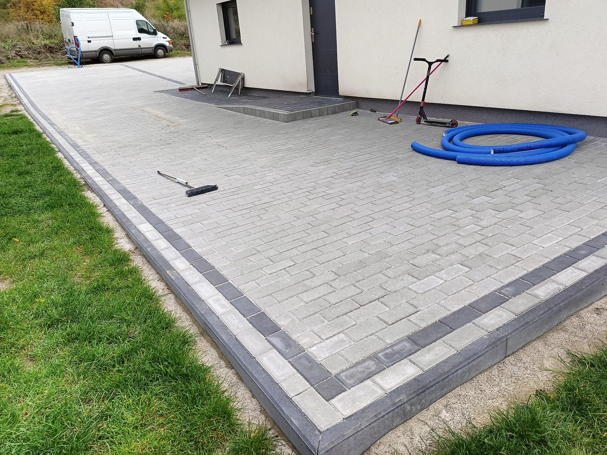 Świeżo ułożona, jasnoszara kostka brukowa z ciemnoszarą obwódką, prowadząca do budynku z ciemnymi drzwiami, na której leżą niebieskie rury drenażowe, a obok stoją hulajnoga i miotła.
