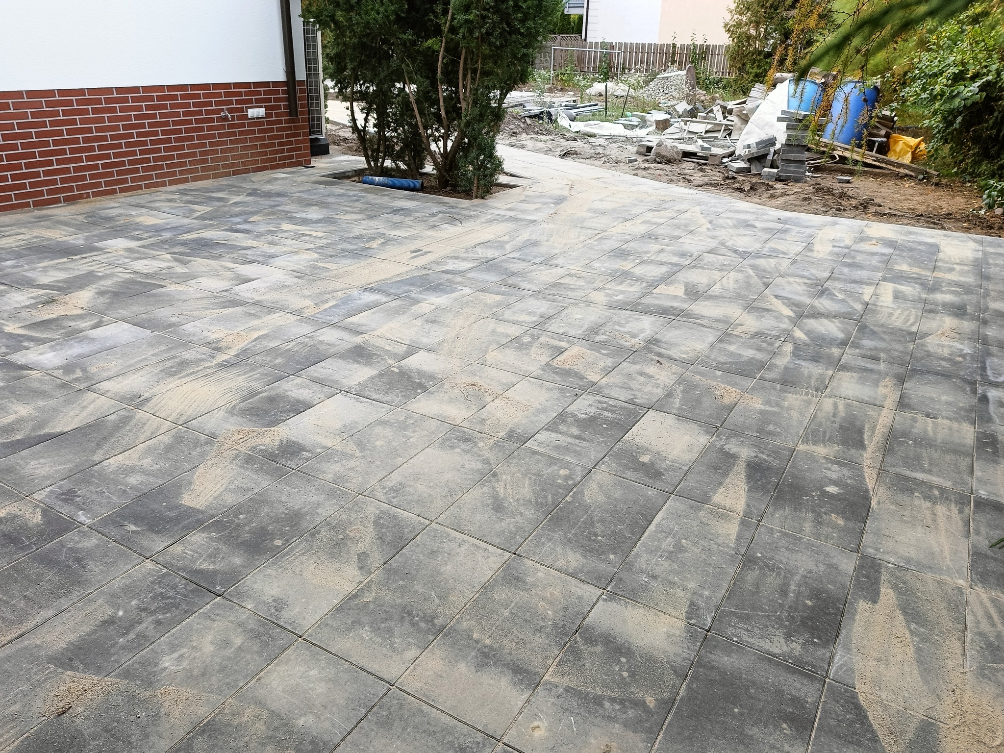 Ułożona szara kostka brukowa na posesji, widoczne ślady piasku na powierzchni, fragment budynku z ceglaną elewacją w tle, teren z przygotowanymi materiałami budowlanymi w oddali.