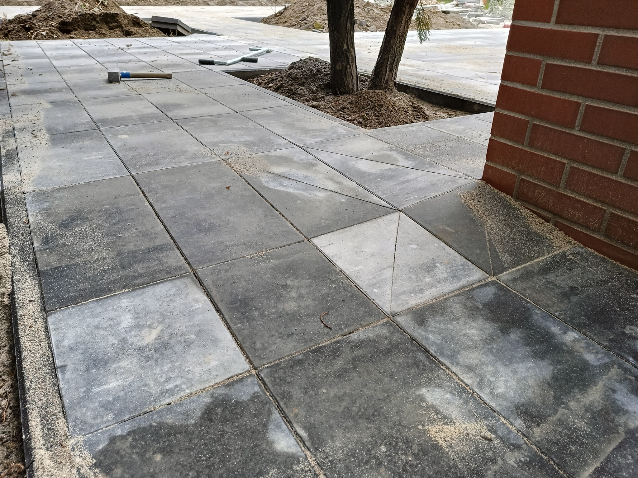 Ułożona szara kostka brukowa wokół drzew, z widocznym młotkiem brukarskim i fragmentem ściany z czerwonej cegły.