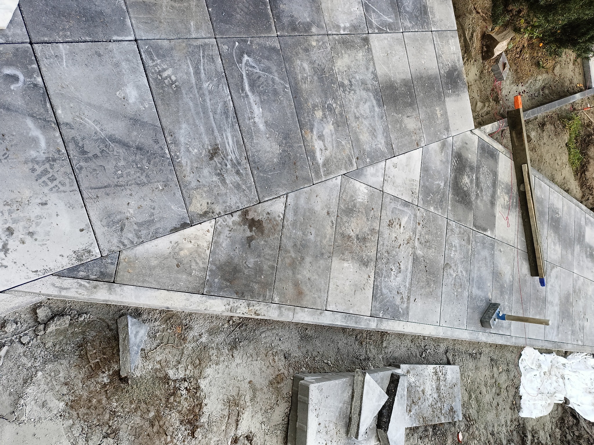 Szare, prostokątne płyty chodnikowe ułożone na piasku, widoczne ślady stóp, narzędzia brukarskie i niezakończony fragment nawierzchni.