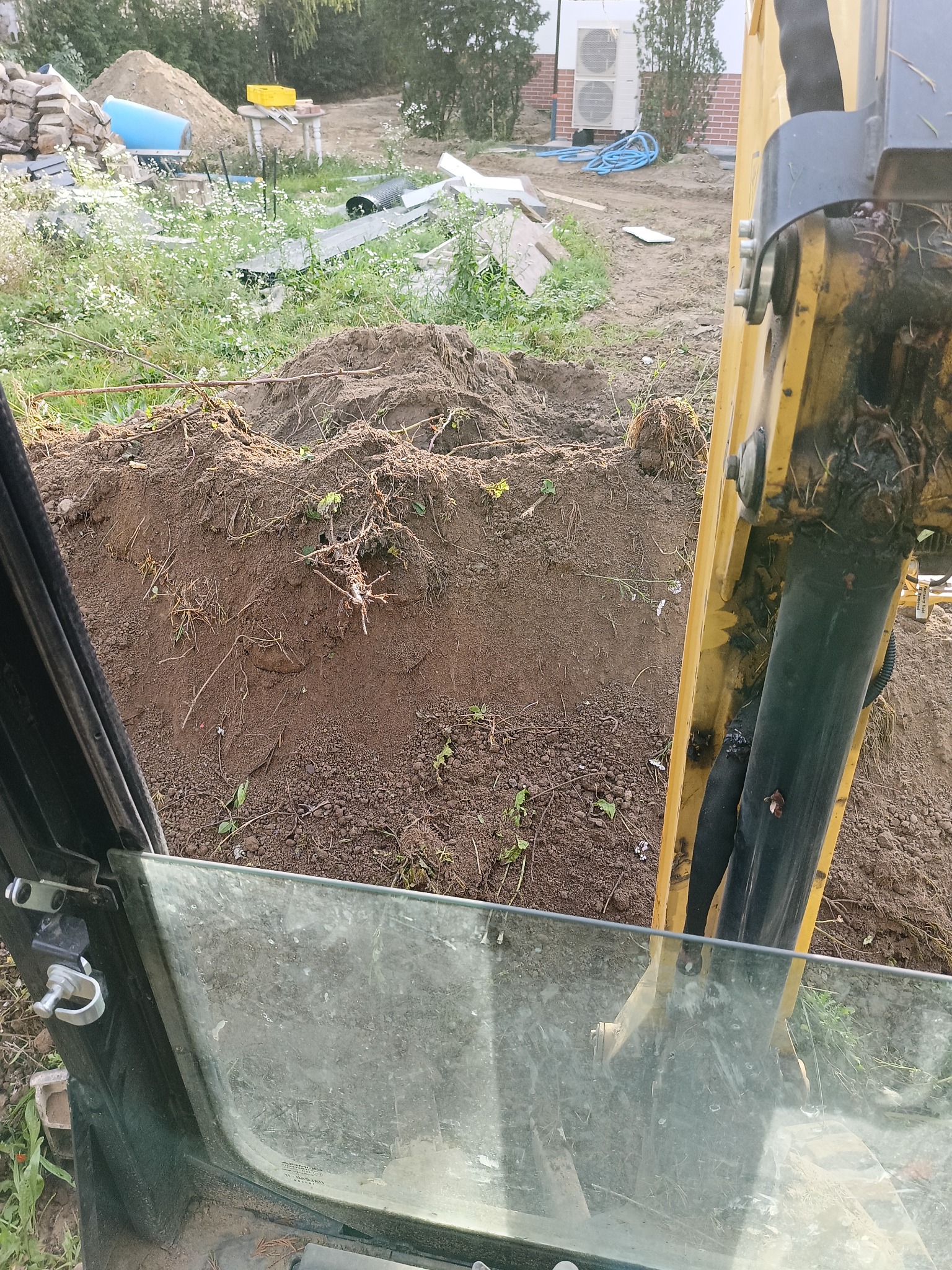 Widok z kabiny koparki na wykopaną ziemię, widoczne ramię maszyny i elementy otoczenia placu budowy, w tle budynek z klimatyzatorami.