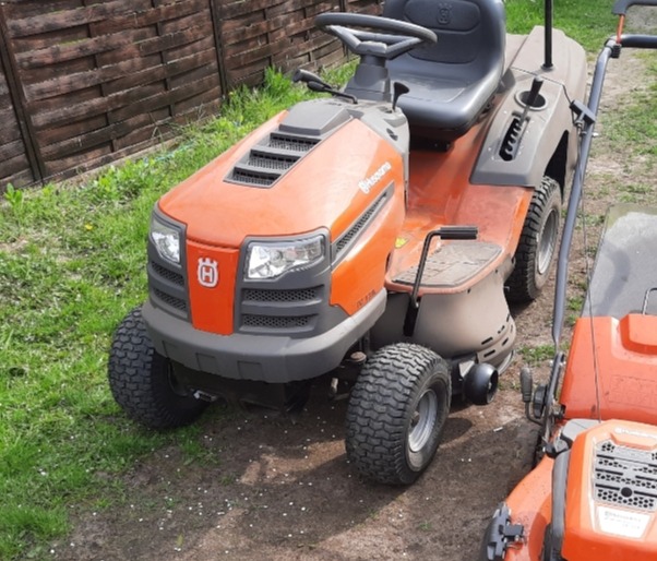 Pomarańczowy traktorek-kosiarka Husqvarna DC110 z widocznym logo na tle trawy i ogrodzenia.