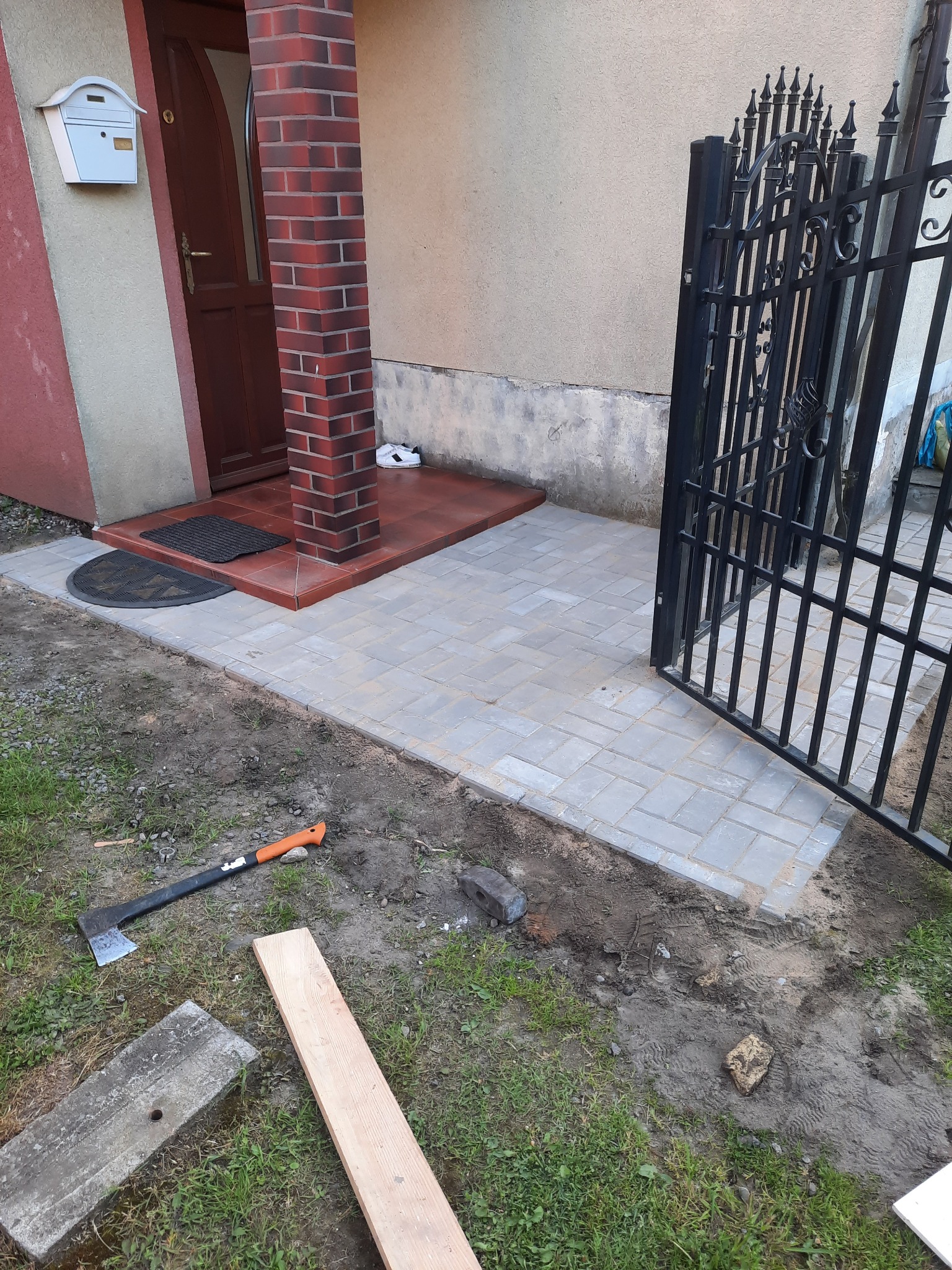 Ułożona kostka brukowa przed domem z widoczną bramką, narzędziami i deskami na trawie.