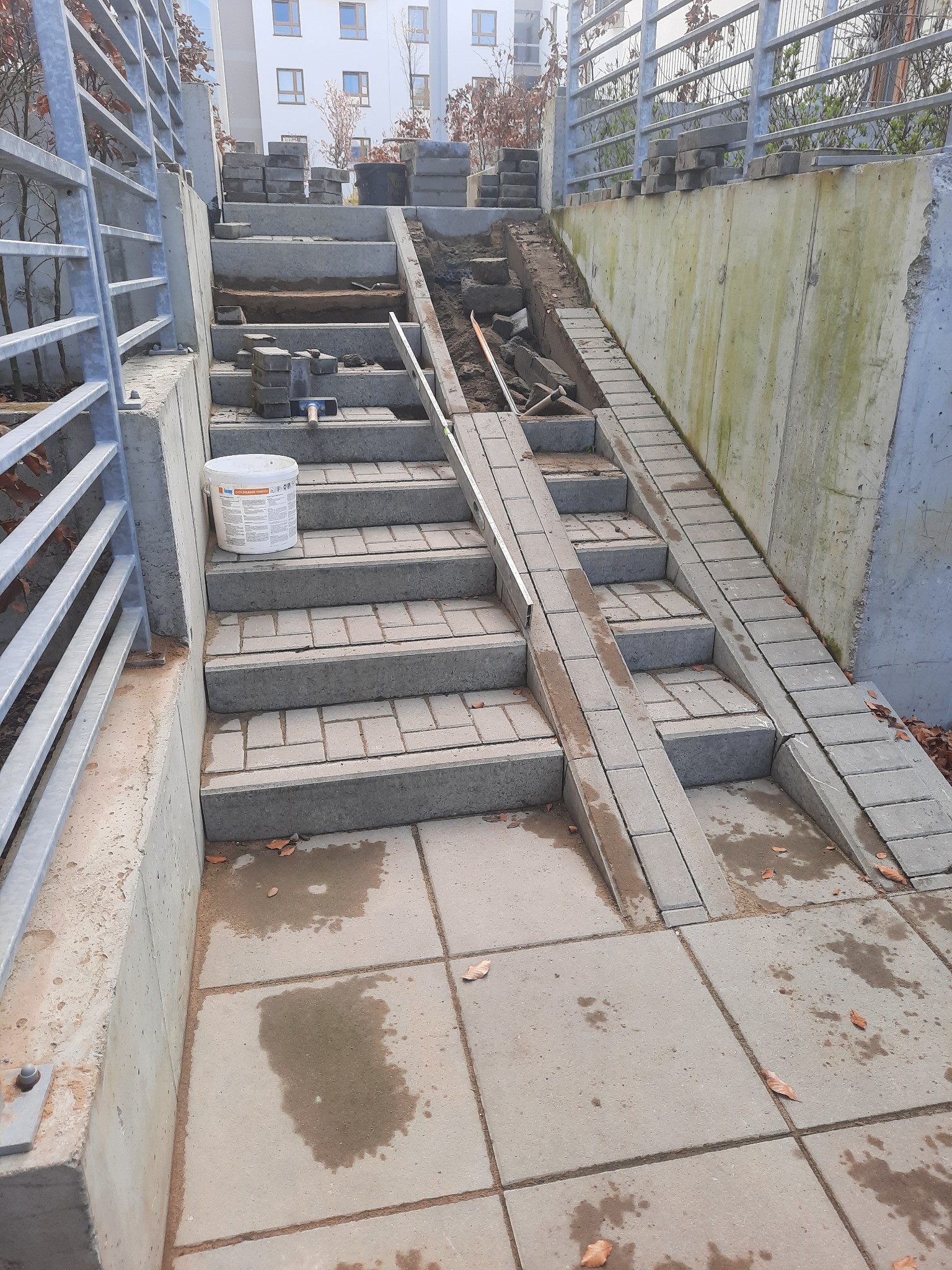 Schody z kostki brukowej w trakcie budowy, widoczne narzędzia, poziomica i wiadro z zaprawą, częściowo ułożona nawierzchnia, tło stanowi budynek mieszkalny.