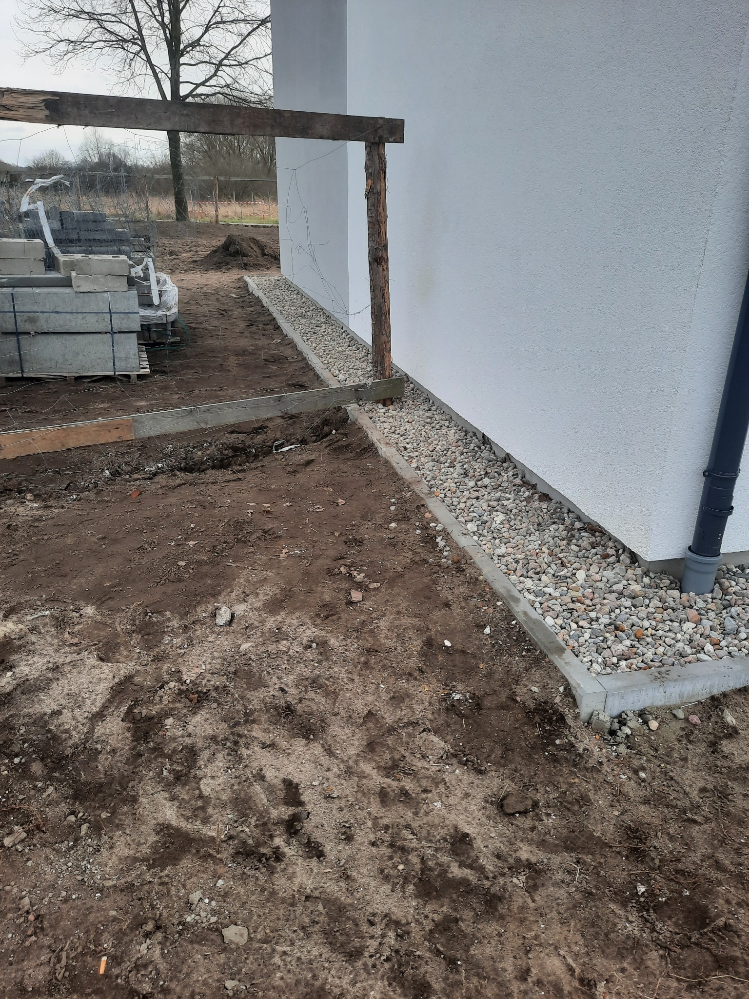 Wykonany drenaż z kamieni obok ściany budynku w Czarnogłowach, widoczny rów wyłożony obrzeżami i wypełniony drobnymi kamieniami, obok rury spustowej.