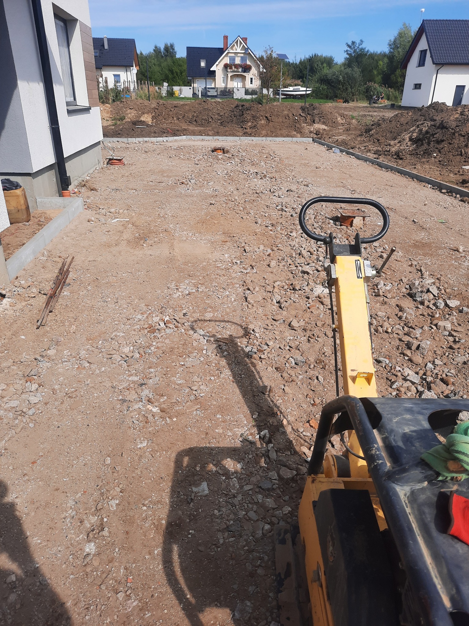 Żółta zagęszczarka wibrująca na gruncie przygotowanym pod budowę z widocznymi obrzeżami, w tle domy jednorodzinne.