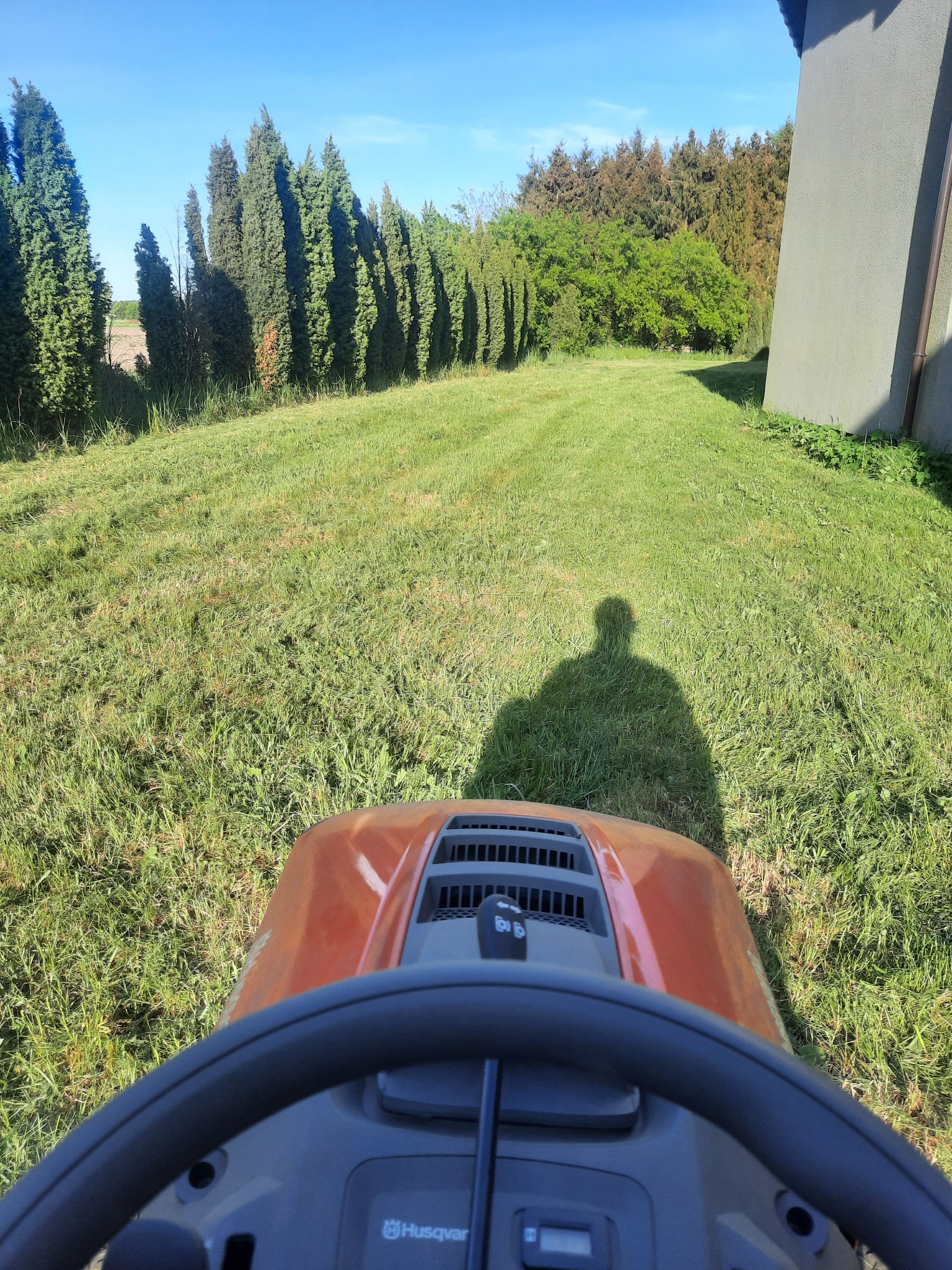 Kosiarka traktorek Husqvarna w trakcie koszenia trawnika, widoczny cień operatora, równo skoszona trawa, rząd drzew w tle, dom po prawej stronie.