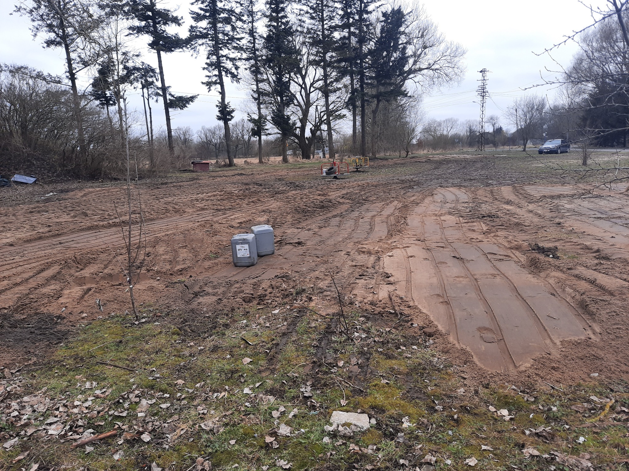 Przygotowany teren pod budowę z widocznymi śladami opon, dwoma pojemnikami i młodymi drzewkami, w tle las i słup wysokiego napięcia.
