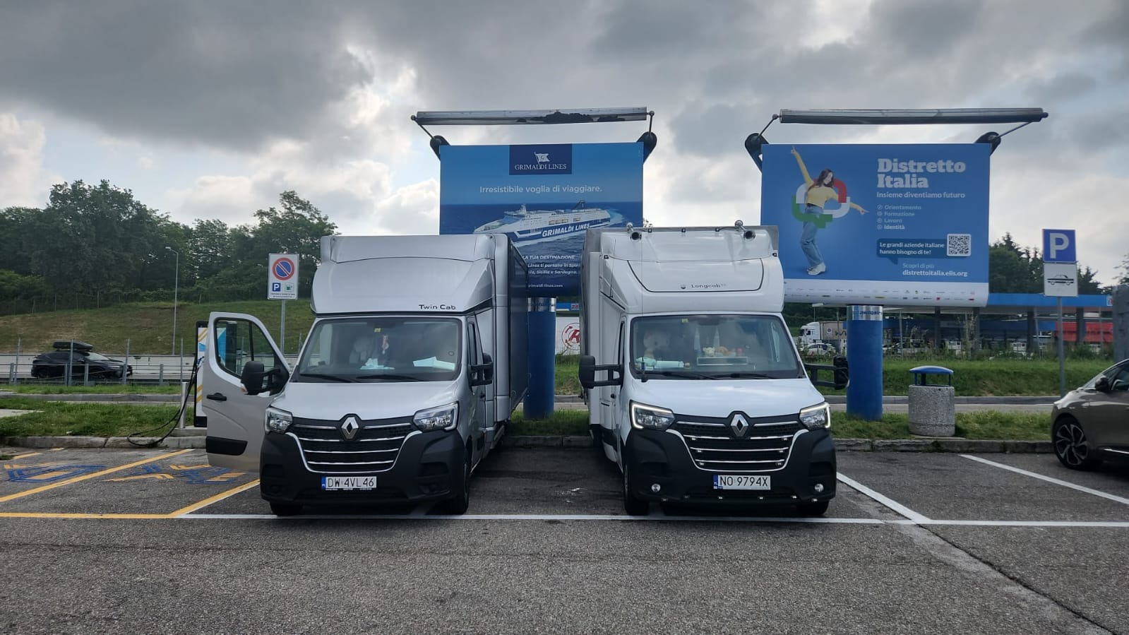 Dwa białe busy Renault Master z wysoką zabudową, zaparkowane obok siebie na parkingu, z reklamami Grimaldi Lines i Distretto Italia w tle. Jeden bus z otwartymi drzwiami.