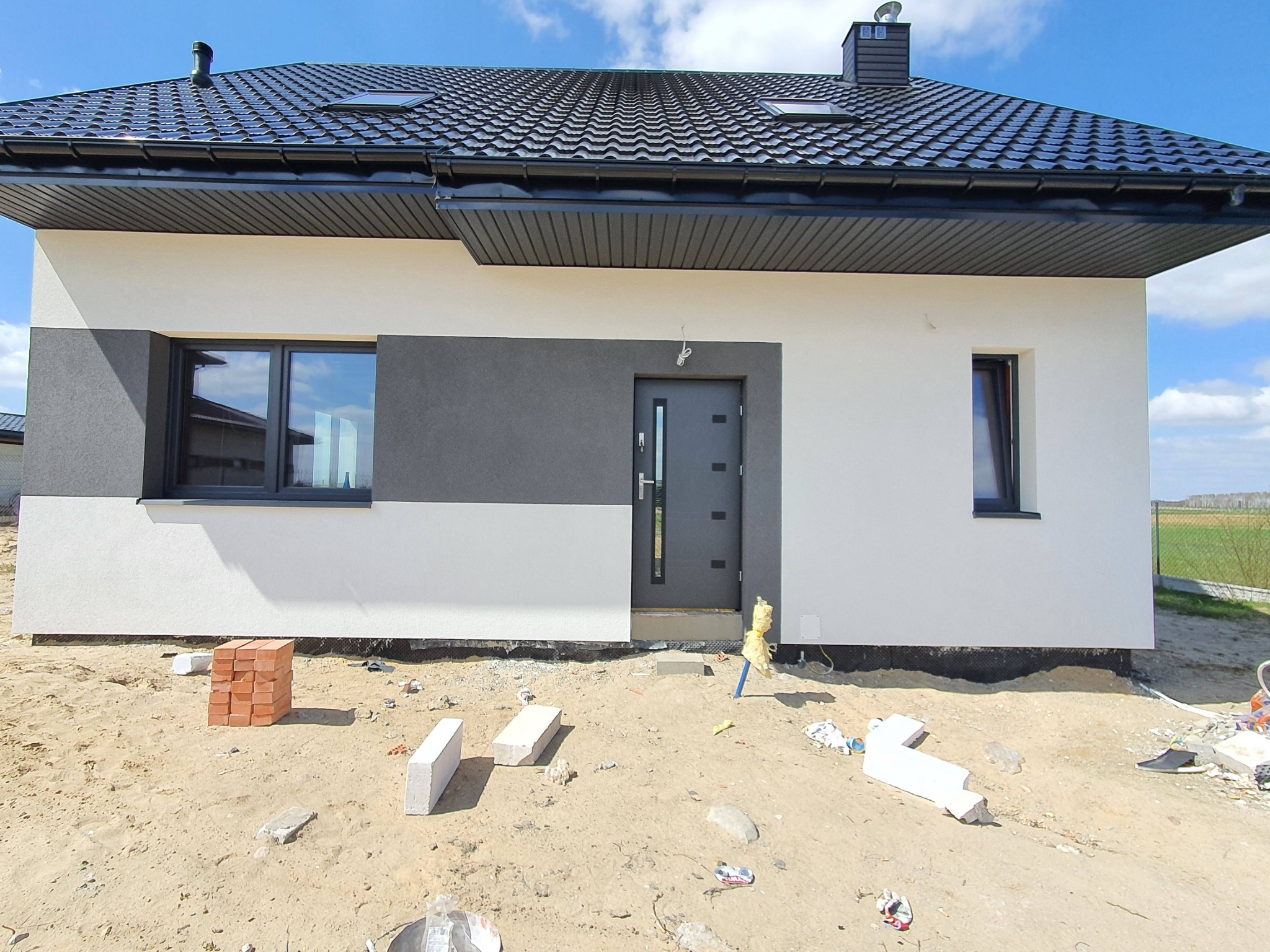 Nowoczesny dom jednorodzinny z elewacją w kolorach bieli i szarości, czarna dachówka, widoczne materiały budowlane przed budynkiem, wskazujące na trwające prace wykończeniowe.