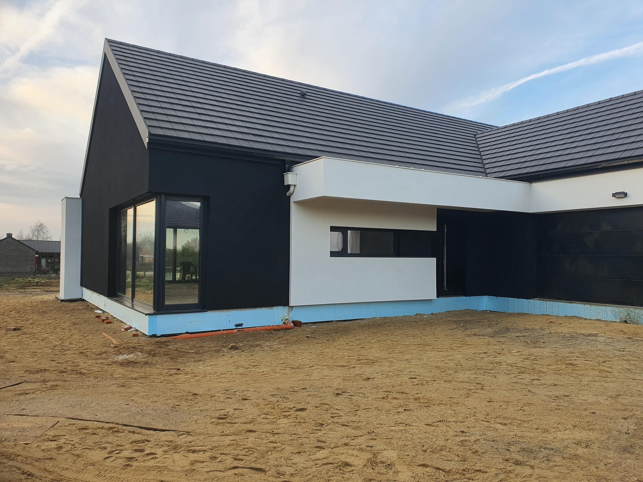 Nowoczesny dom jednorodzinny z czarną elewacją i białymi elementami architektonicznymi, widoczna warstwa izolacji fundamentów oraz teren przygotowany pod zagospodarowanie.