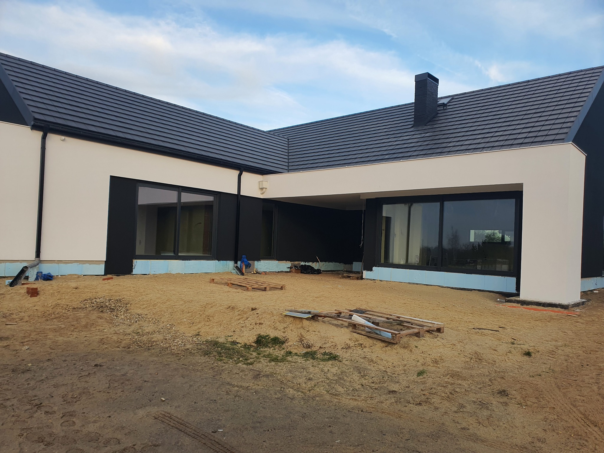 Nowoczesny dom jednorodzinny w trakcie prac wykończeniowych elewacji, widoczne styropianowe płyty izolacyjne przy fundamencie, teren wokół domu częściowo wyrównany piaskiem, palety budowlane...