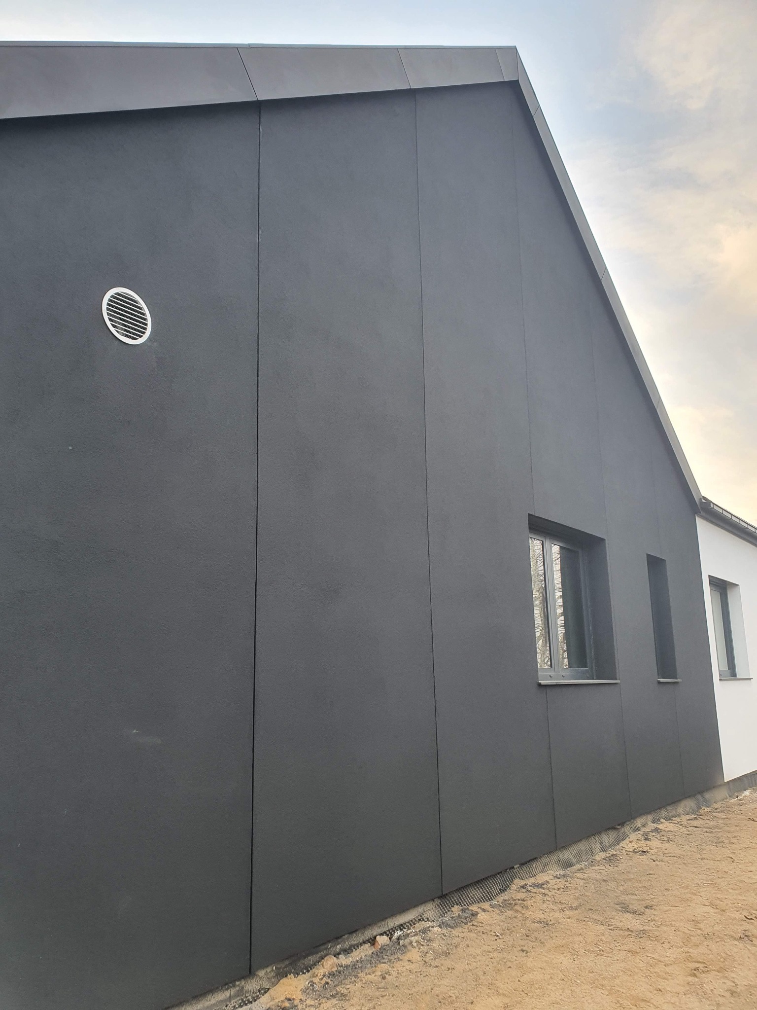 Nowoczesna elewacja domu jednorodzinnego w kolorze grafitowym, z widoczną wentylacją i dwoma oknami o różnych rozmiarach, wykończona panelami, teren wokół domu w trakcie prac ziemnych.