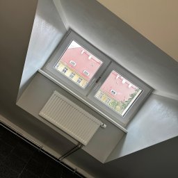 USŁUGI REMONTOWO-BUDOWLANE KAMILA KOTKOWSKA - Poddasze po szpachlowaniu i malowaniu na szaro, z widocznym oknem dachowym, grzejnikiem i fragmentem czarnej podłogi.