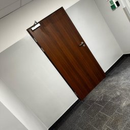 USŁUGI REMONTOWO-BUDOWLANE KAMILA KOTKOWSKA - Ciemnobrązowe drzwi z chromowanymi okuciami w białym pomieszczeniu z szarą podłogą z płytek. Widoczny samozamykacz na górze drzwi i czujnik bezpieczeństwa na ścianie.