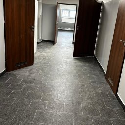 USŁUGI REMONTOWO-BUDOWLANE KAMILA KOTKOWSKA - Korytarz biurowy z szarą terakotą na podłodze i drewnianymi drzwiami, widoczne niedociągnięcia w suficie podwieszanym.
