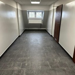 USŁUGI REMONTOWO-BUDOWLANE KAMILA KOTKOWSKA - Wnętrze korytarza z szarą podłogą z płytek, biało-szare ściany z oknem na końcu i brązowe drzwi po prawej stronie.