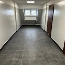 Wnętrze korytarza z szarą podłogą z płytek, biało-szare ściany z oknem na końcu i brązowe drzwi po prawej stronie.