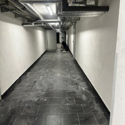 USŁUGI REMONTOWO-BUDOWLANE KAMILA KOTKOWSKA - Korytarz z ciemną, prostokątną posadzką w trakcie czyszczenia, widoczne rury pod sufitem i osoba pracująca w oddali.