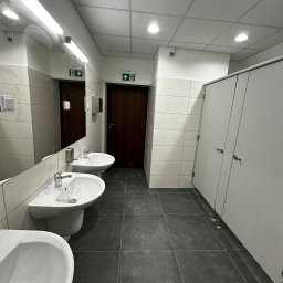 USŁUGI REMONTOWO-BUDOWLANE KAMILA KOTKOWSKA - Wnętrze publicznej toalety z trzema umywalkami, lustrem, płytkami na ścianach i podłodze oraz kabinami WC z białymi drzwiami.
