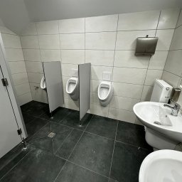 USŁUGI REMONTOWO-BUDOWLANE KAMILA KOTKOWSKA - Wnętrze toalety publicznej z trzema pisuarami oddzielonymi przegrodami, umywalką i dozownikiem ręczników na ścianie wyłożonej jasnymi płytkami, podłoga z ciemnych płytek.
