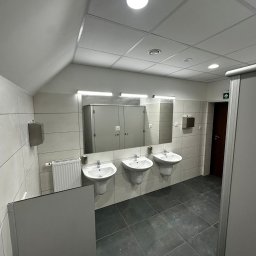 USŁUGI REMONTOWO-BUDOWLANE KAMILA KOTKOWSKA - Wnętrze publicznej toalety z trzema umywalkami na postumentach, dużym lustrem z oświetleniem LED, szarymi płytkami ściennymi i podłogowymi, oraz kabinami WC z drzwiami.