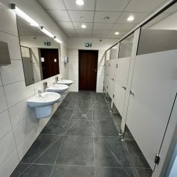 USŁUGI REMONTOWO-BUDOWLANE KAMILA KOTKOWSKA - Wnętrze publicznej toalety z trzema umywalkami, dużym lustrem z oświetleniem, kabinami WC z szarymi drzwiami i ciemną podłogą z płytek. Nad drzwiami widoczne znaki ewakuacyjne.